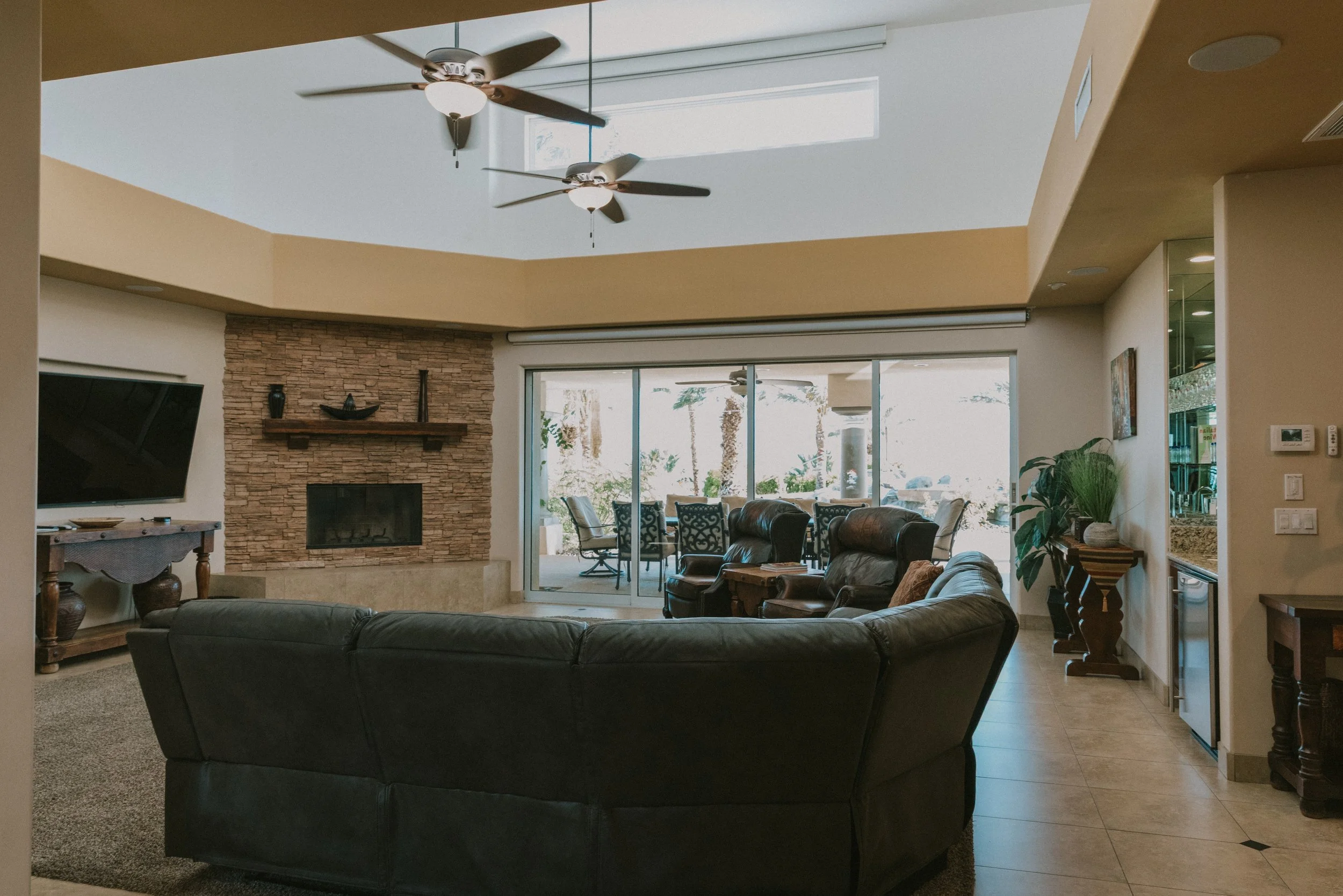 Living room with a black leather sofa, stone fireplace, ceiling fans with lights, sliding glass doors with potted plant and outdoor patio furniture visible outside.