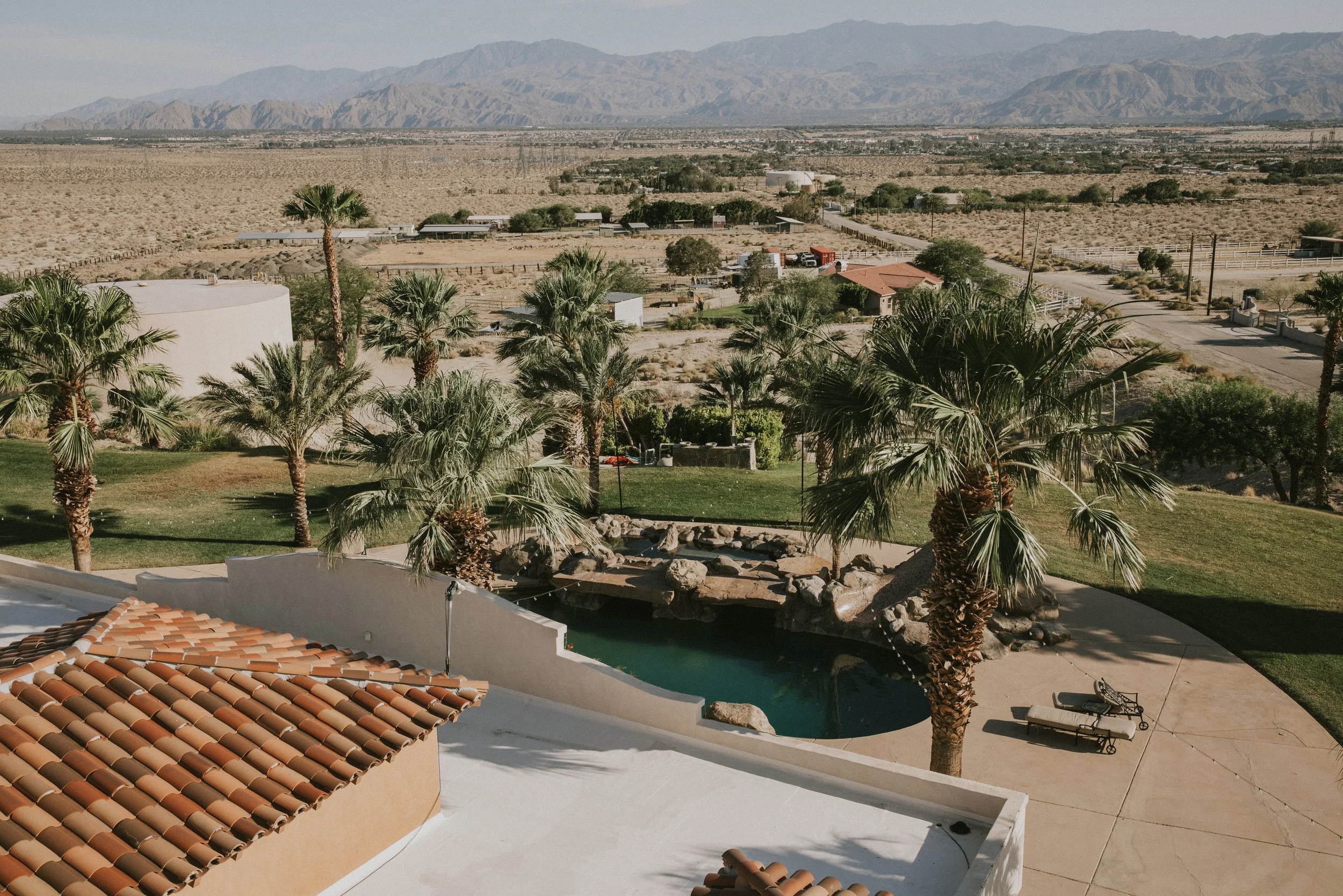 View of a desert landscape with mountains in the background, palm trees, a swimming pool, and a patio area with lounge chairs under a clear sky.