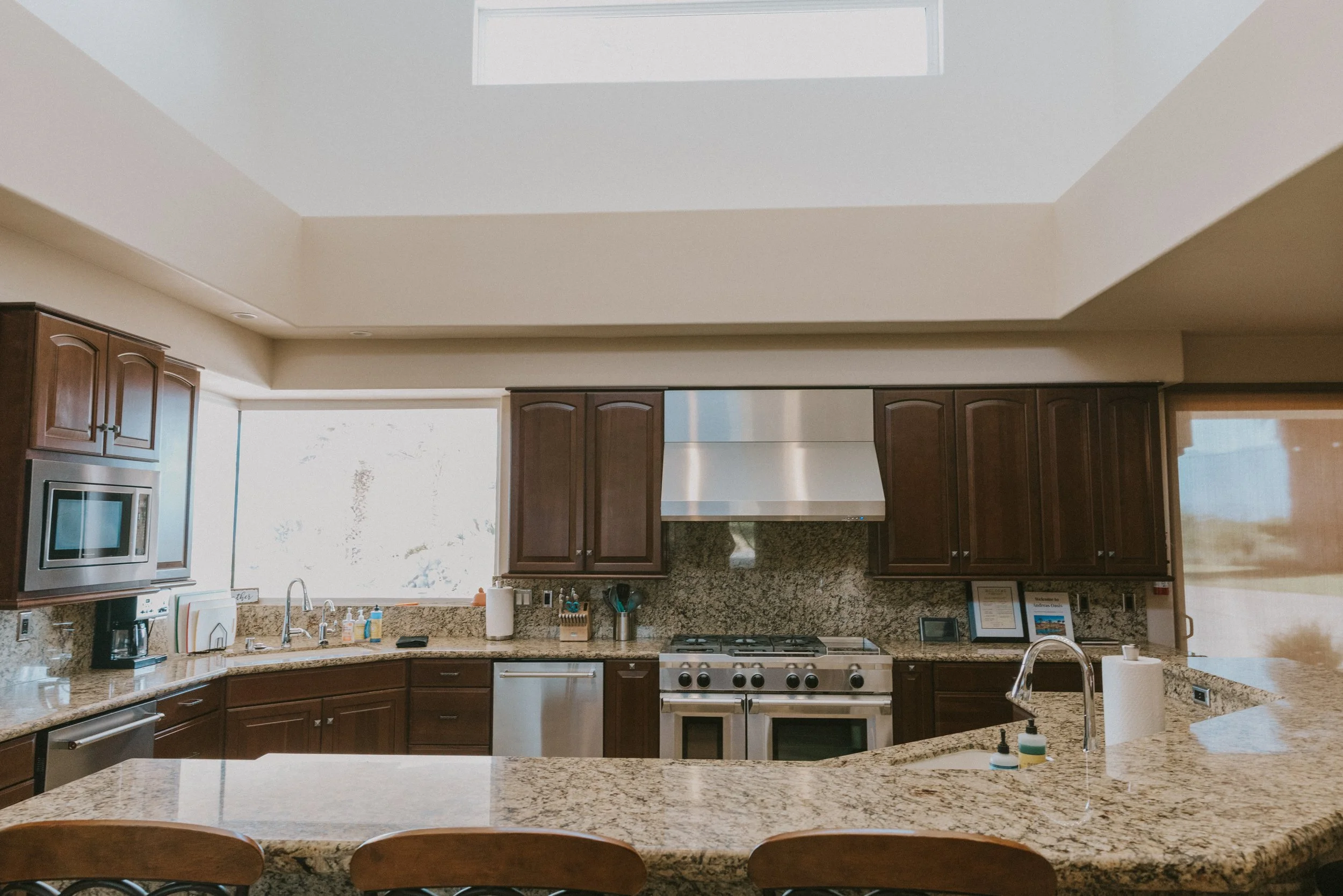 A kitchen with brown wooden cabinets, granite countertops, stainless steel appliances, a stove, a microwave, and a double sink. There are paper towels, soap, and various kitchen items on the counter. A large window and a skylight illuminate the space