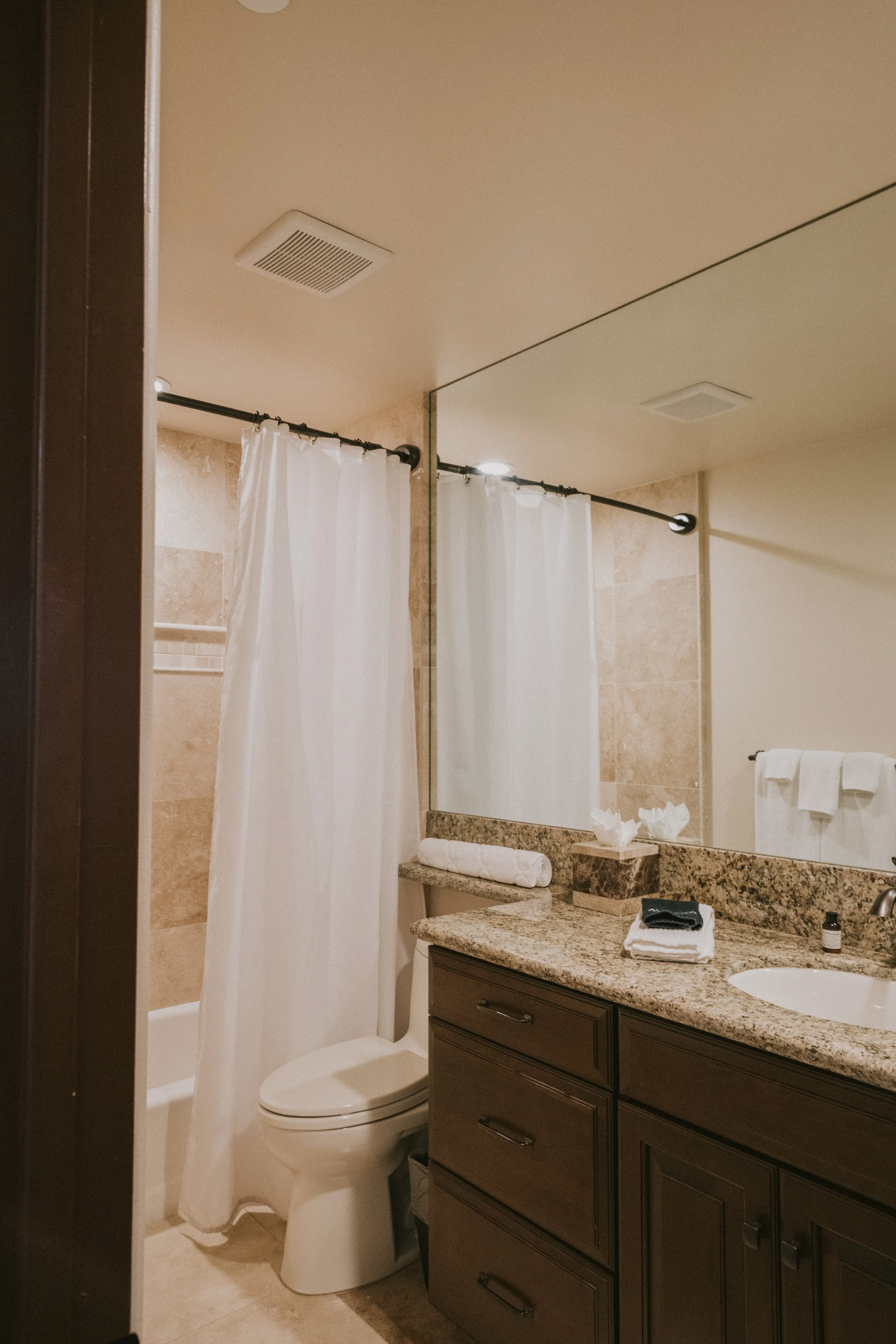A bathroom with a toilet, a granite countertop with a sink and folded towels, a mirror, and a shower with white curtain rod and curtain. The ceiling has two ventilation fans and a light.