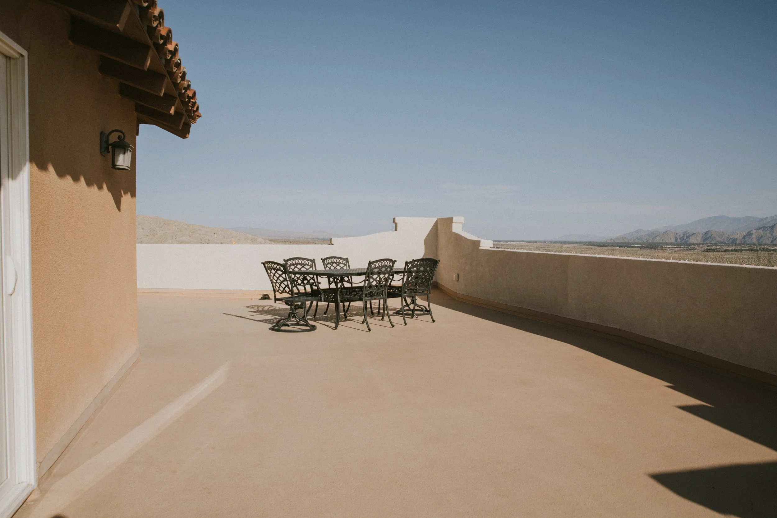 A spacious outdoor terrace with a round metal table and matching chairs, overlooking a desert landscape with mountains in the distance, under a clear blue sky.