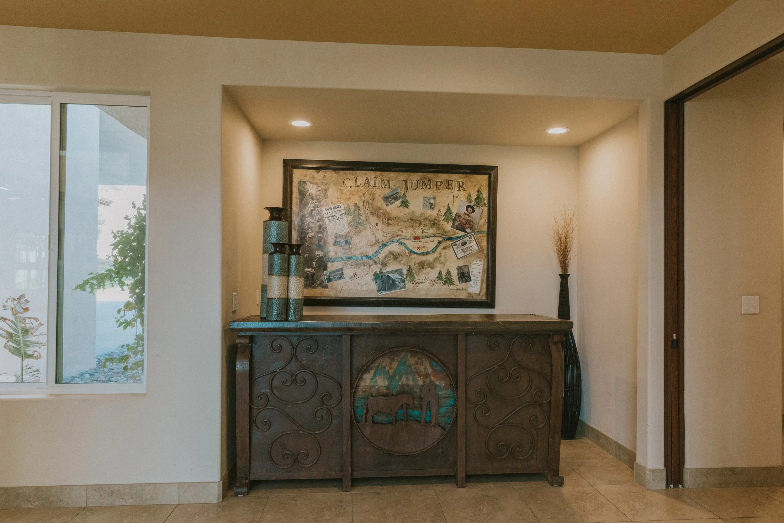 Interior view of a room with a framed map titled 'Claim Jumper,' a wooden console table, tall decorative vases, and a window showing greenery outside. Recessed ceiling lights illuminate the space.