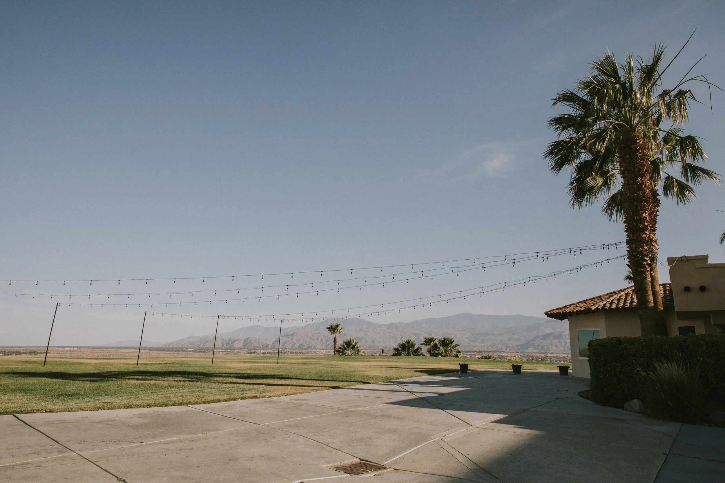 A landscape featuring a clear sky, a palm tree, and a building with a tiled roof. String lights are hanging across the scene, with mountains visible in the background. There is a concrete patio in the foreground.