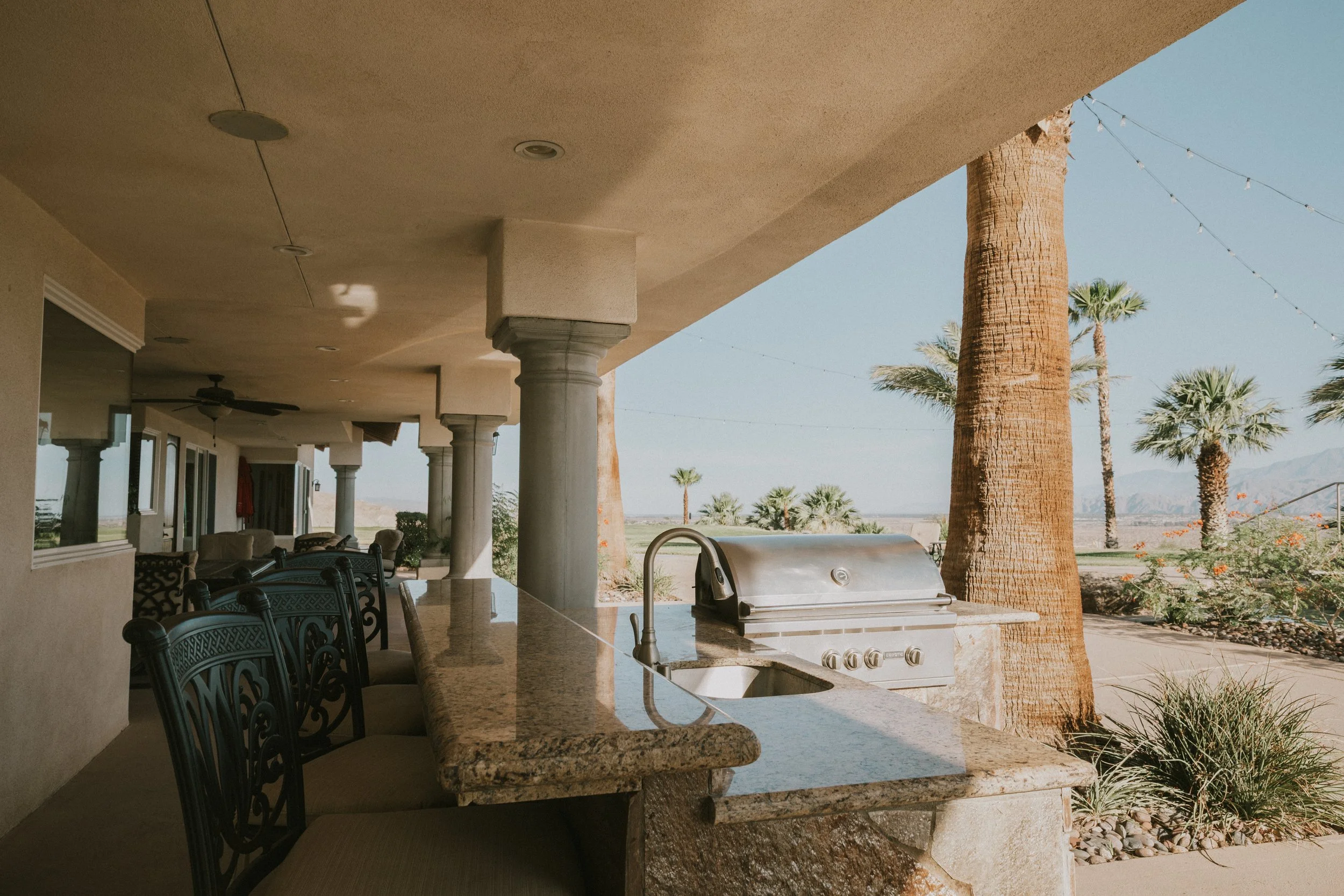 Outdoor patio with a built-in grill, chairs, tables, palm trees, and string lights, overlooking a desert landscape.