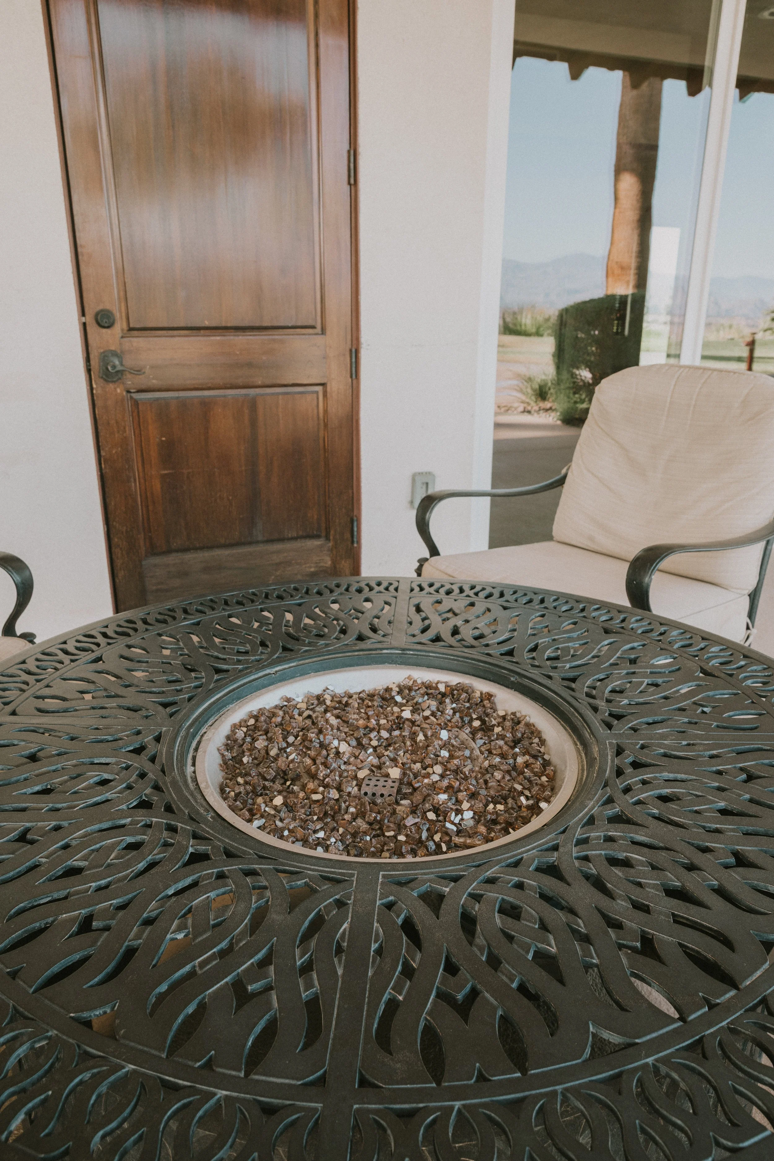 A round outdoor fire pit filled with small decorative stones, with a decorative metal table surrounding it, situated on a patio near a house with a wooden door and large glass windows showing an outdoor landscape with mountains.