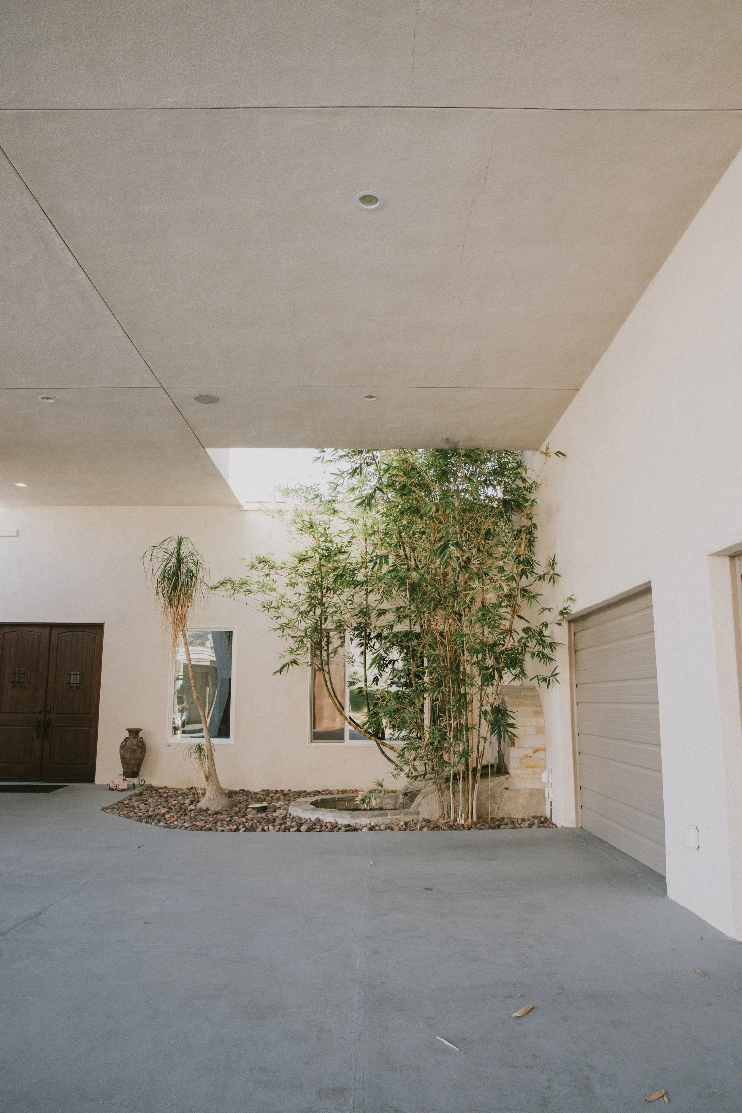 A modern house entrance with a concrete driveway, a garage door, a wooden door with a small window, decorative vases, and a garden with trees and rocks.