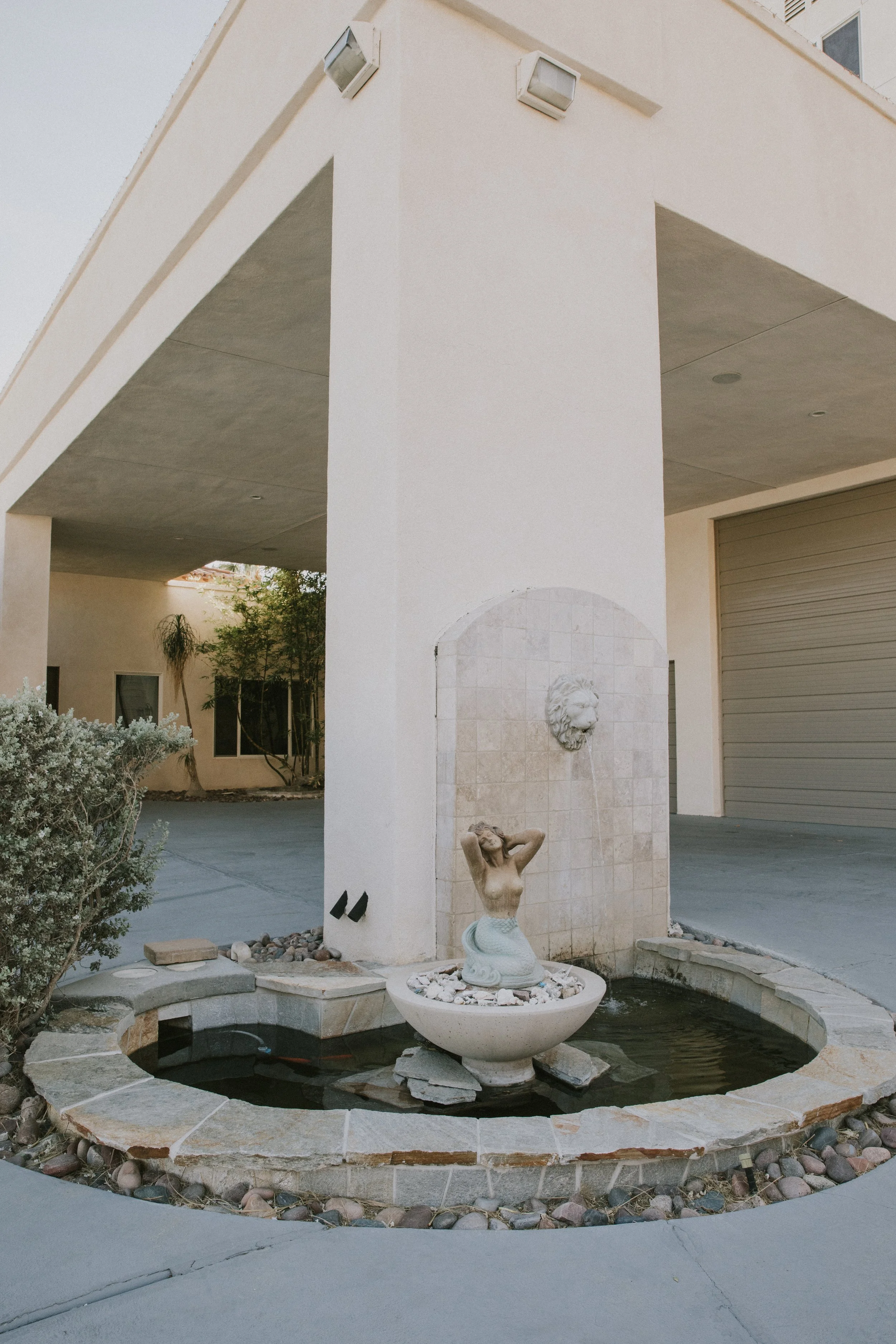 A courtyard area with a small fountain featuring a mermaid sculpture, surrounded by a stone border and rocks. A white wall with a lion face water spout is behind the fountain, and there is a large garage door and house windows in the background.