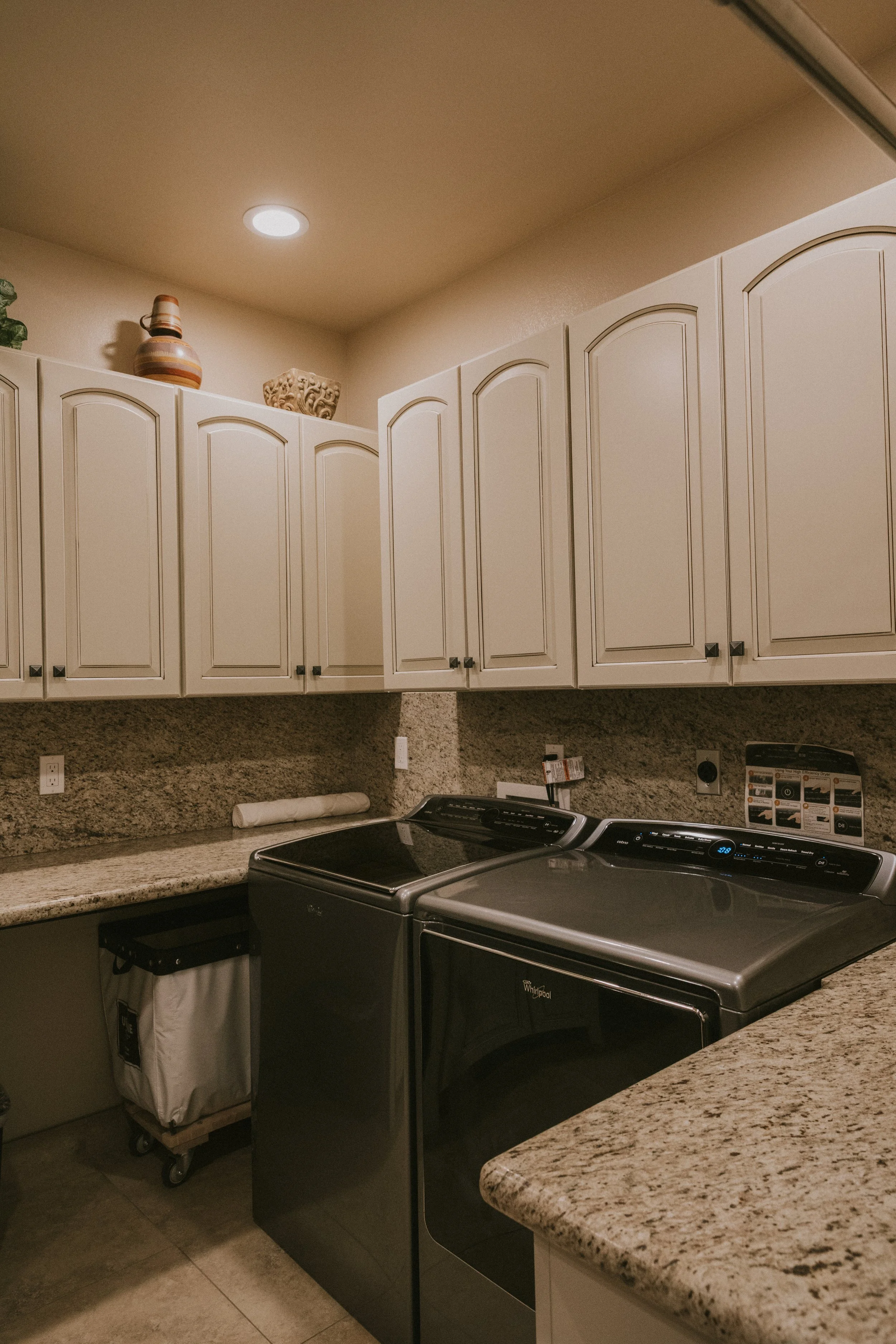 Kitchen with white cabinets, granite countertops, and a Whirlpool washing machine. Items on top include decorative vases and a basket. A paper towel roll is on the counter, and a laundry basket with a cover is beneath the counter.