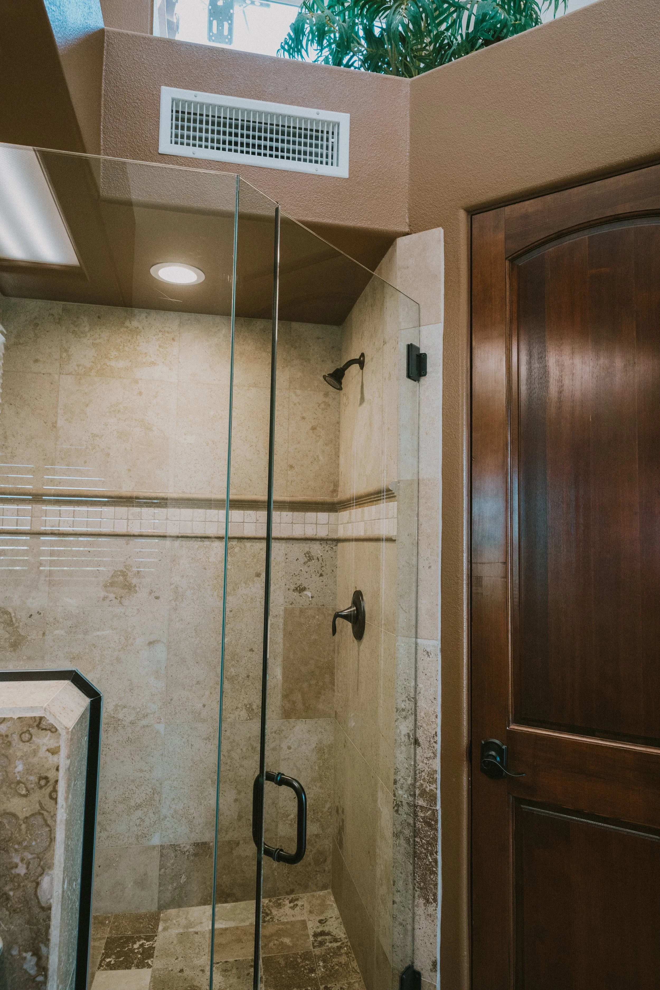 A bathroom shower area with beige tiles, a glass door, a black showerhead, and a wooden cabinet door to the right. There is a ceiling vent and a window with a plant visible outside.