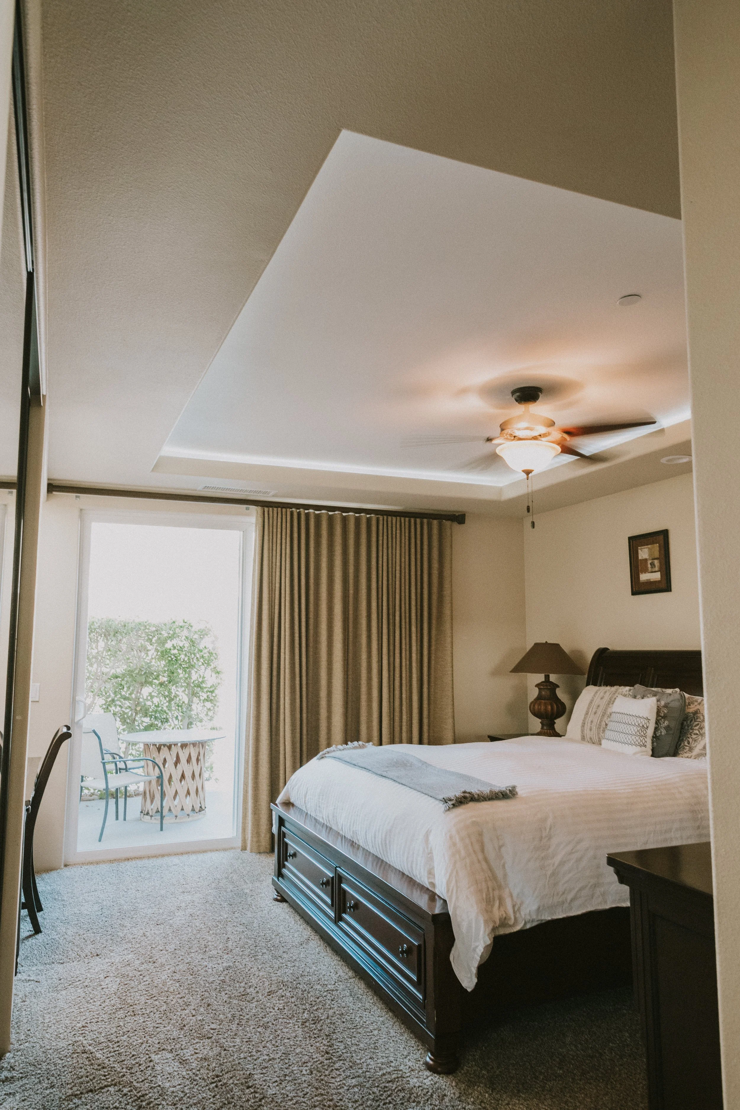 A bedroom with a large bed, nightstand with lamp, and a ceiling fan with light. There's a sliding glass door with curtains leading to an outdoor balcony with two chairs and a small table. The room has beige walls, a framed picture, and a carpeted flo