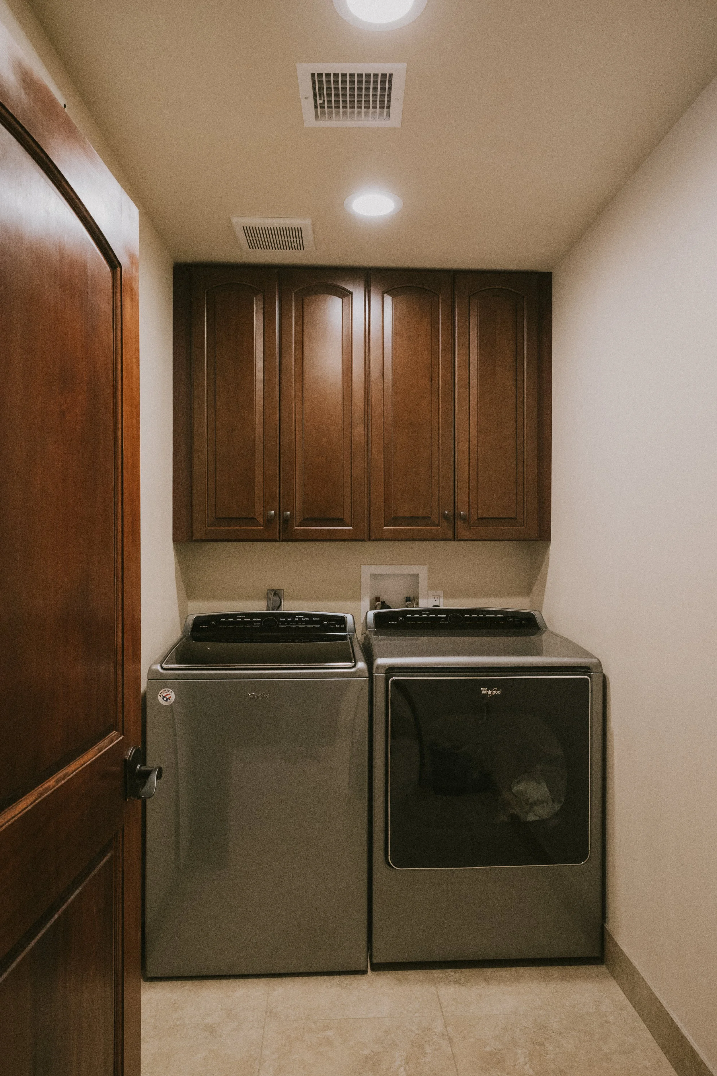 A laundry room with two gray washing machines side by side beneath dark wooden cabinets, with a beige wall and tiled floor.