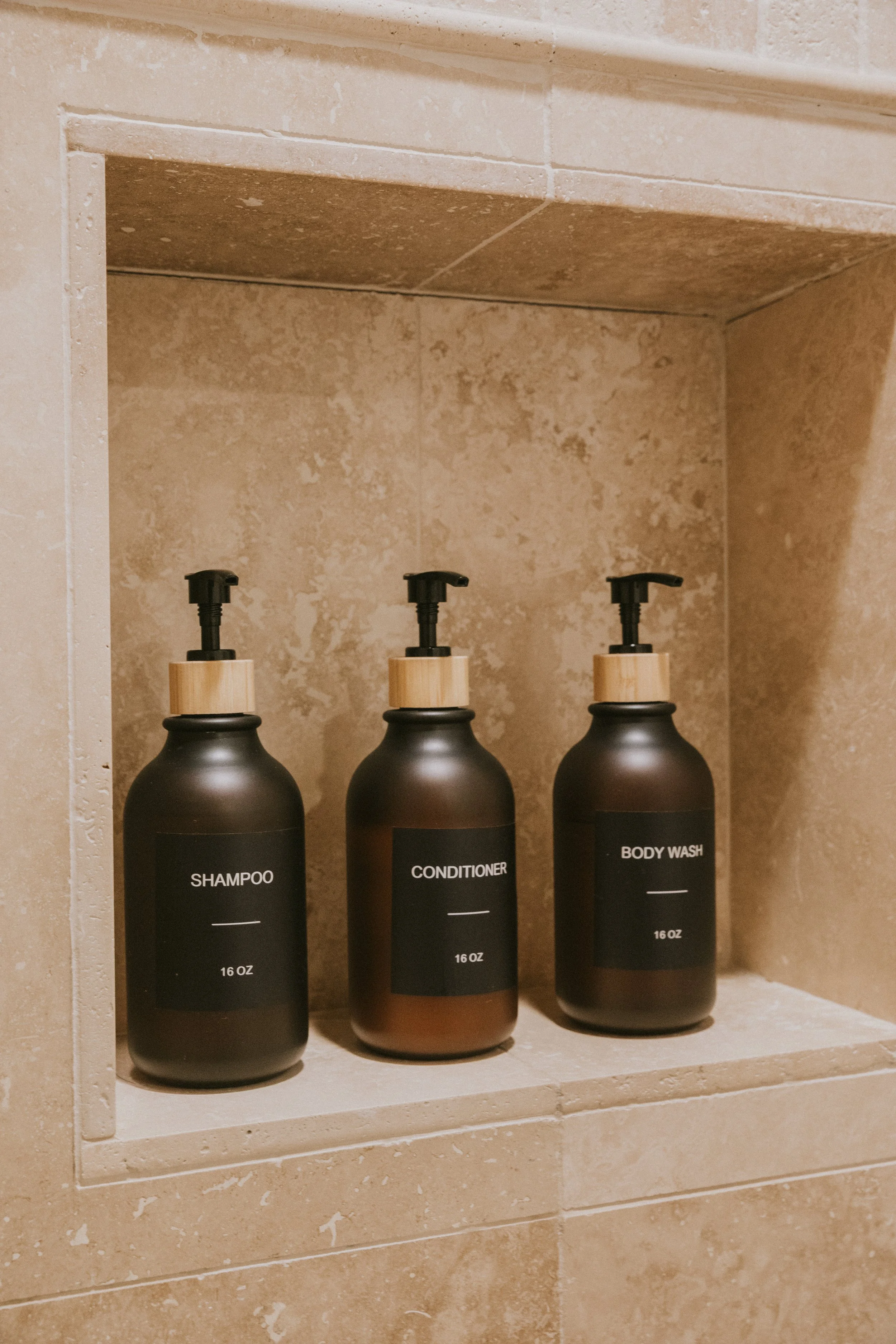 Three black bottles with beige pump dispensers labeled Shampoo, Conditioner, and Body Wash, placed on a beige marble shelf in a bathroom.