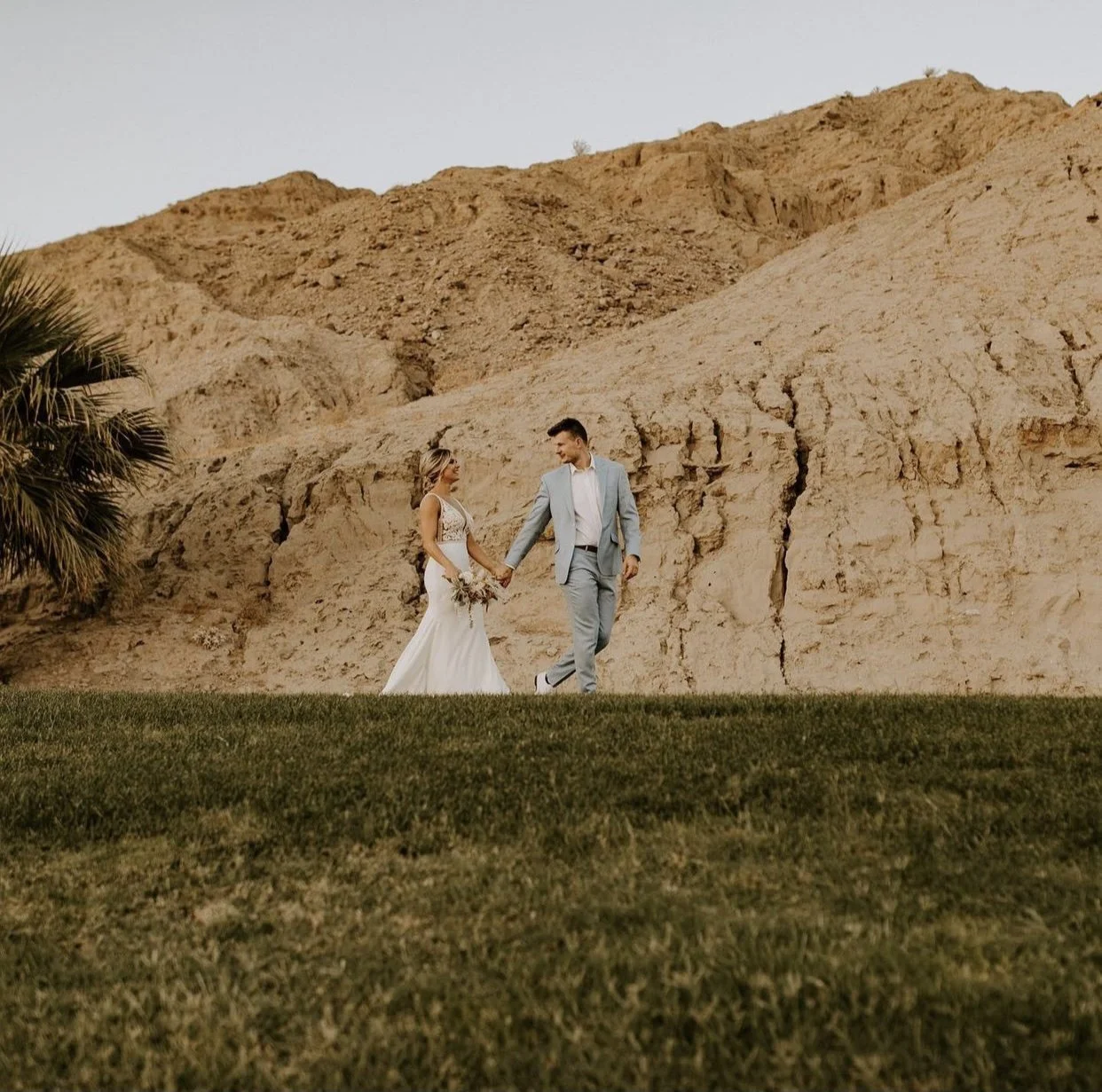 A bride and groom holding hands walking outdoors in front of a rocky cliff, with green grass in the foreground and a palm tree on the left.