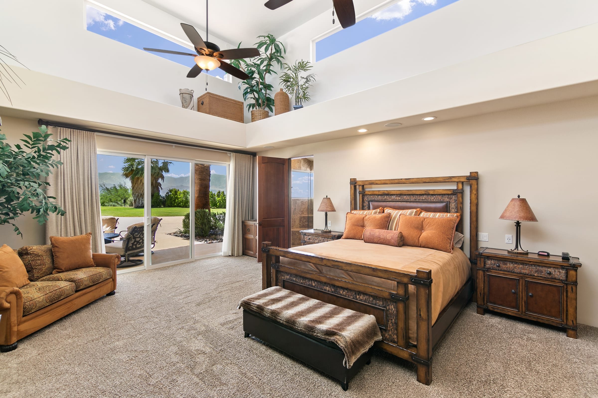 Bright bedroom with a large wooden bed, nightstands with lamps, a beige sofa, and glass sliding doors leading to an outdoor patio with chairs, palm trees, and mountain views.