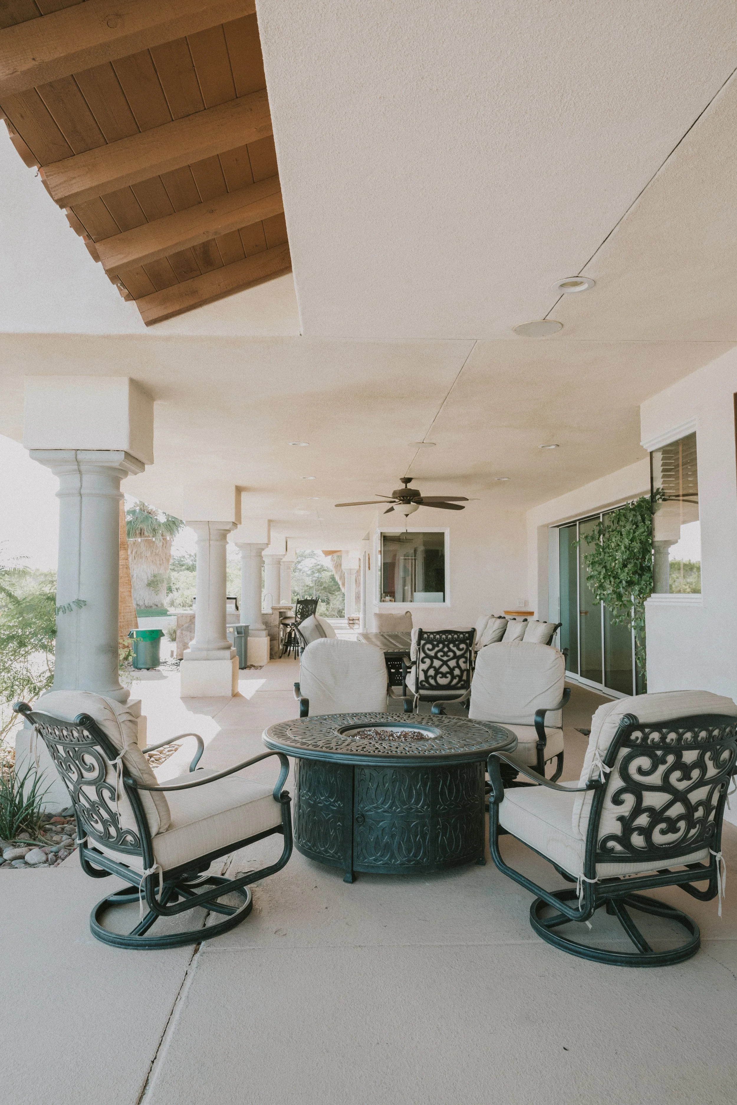 An outdoor patio area with cream-colored cushioned chairs around a decorative fire pit table, supported by elegant black metal frames. The patio has tall columns and a ceiling with recessed lights and a fan. There are sliding glass doors and a view o