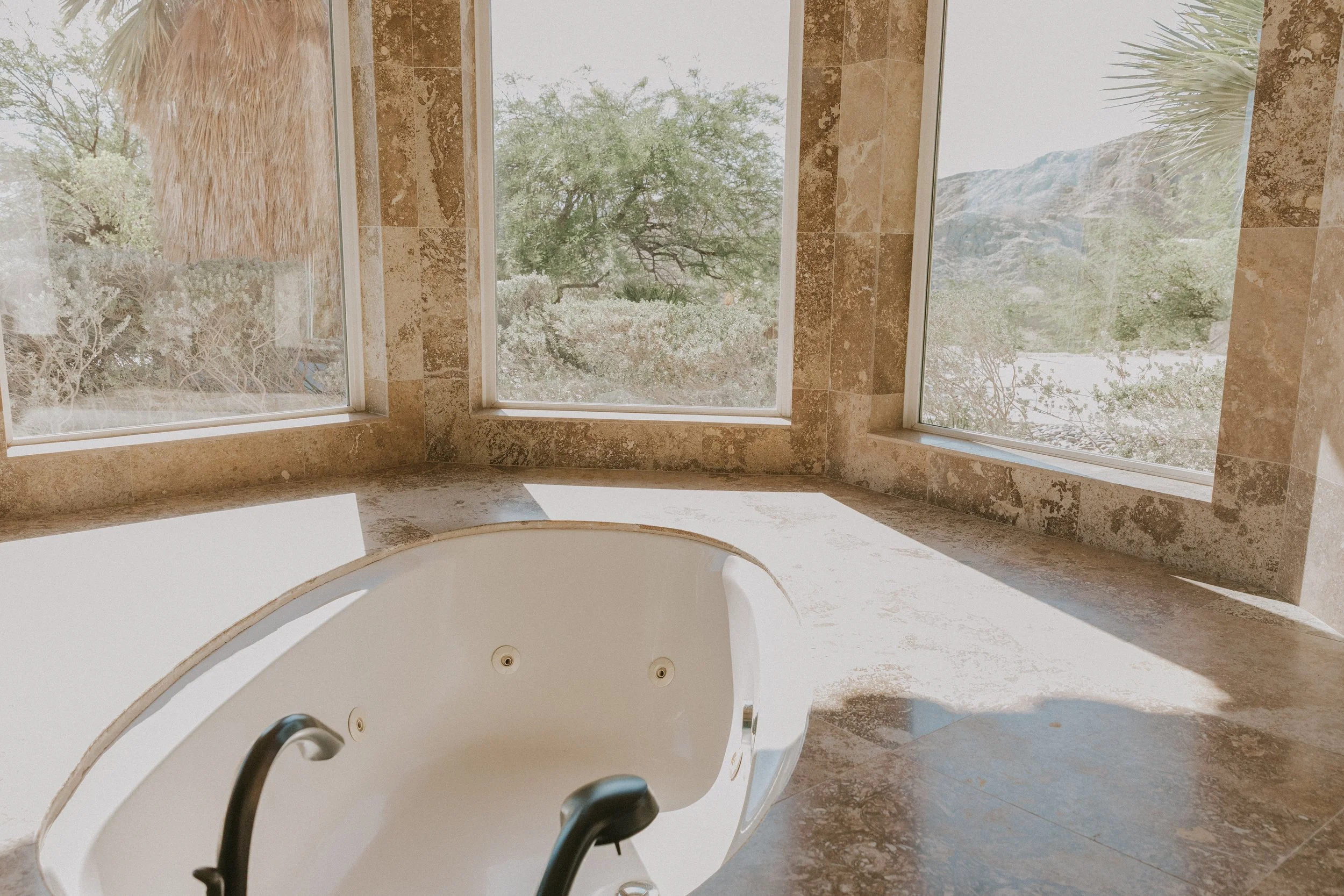 Bathroom with large windows showing trees and outdoor landscape, corner of a bathtub, and marble walls.