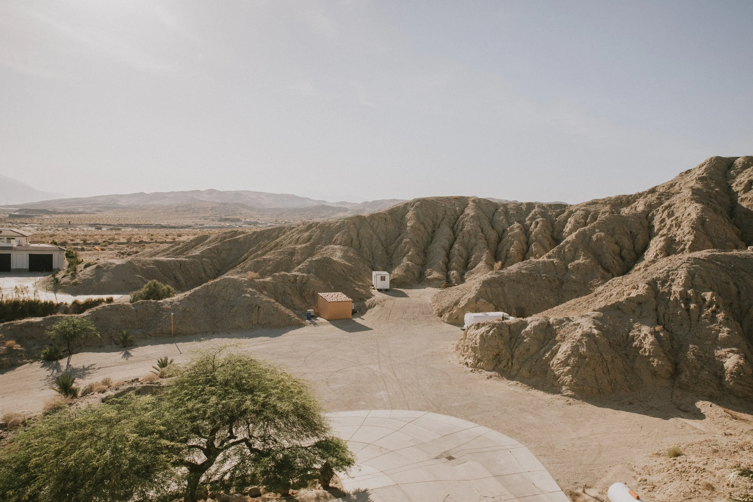 Desert landscape with rugged hills and minimal vegetation, scattered houses, and a dirt road, under a clear sky.