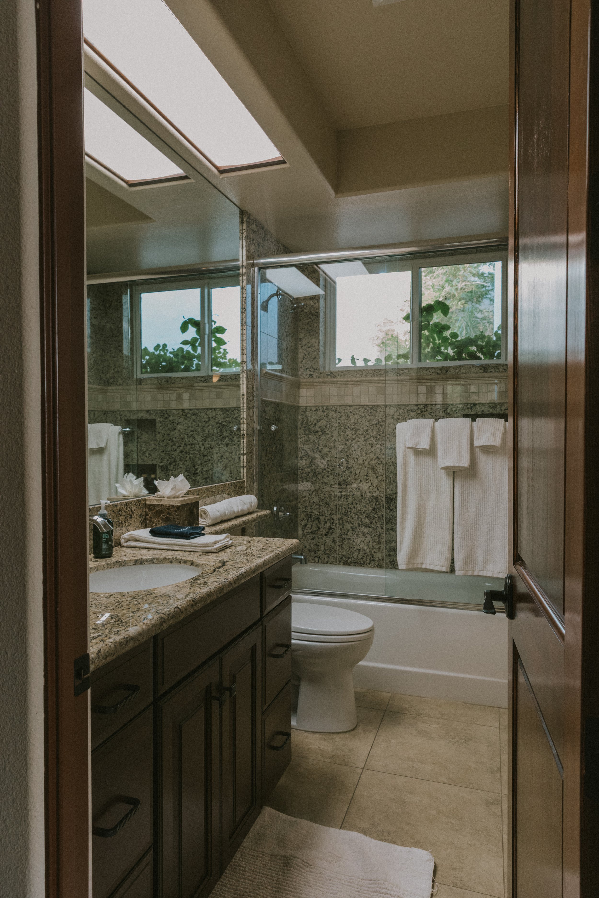 A bathroom with granite countertop, dark wooden vanity, mirror, windows with greenery outside, bathtub with glass door, toilet, towels, and toiletries.