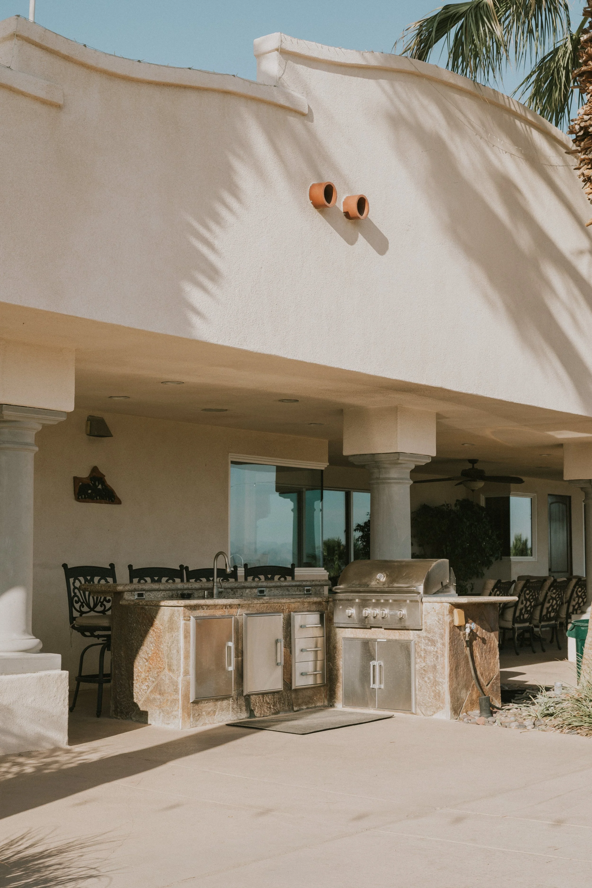 Outdoor patio area with built-in grill, stone countertop, black chairs, and palm trees in the background, under a sunny sky.