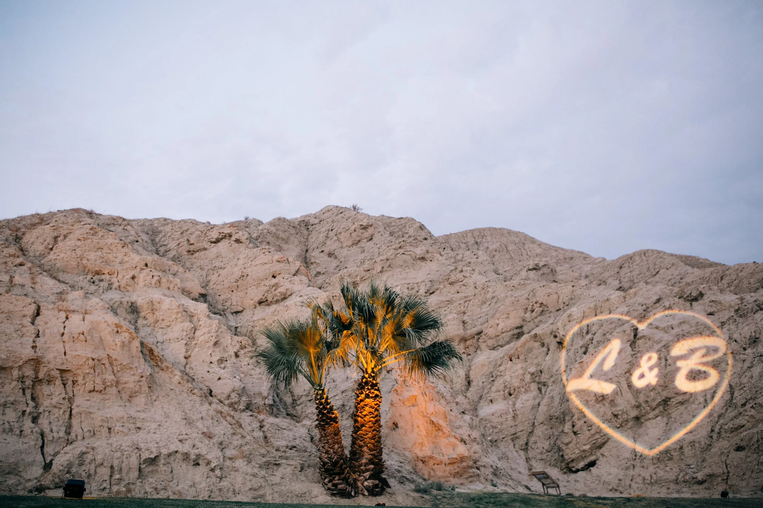 Two palm trees illuminated by yellow lights grow in front of a rocky mountain with a heart shape drawn on it containing the text "L & B".