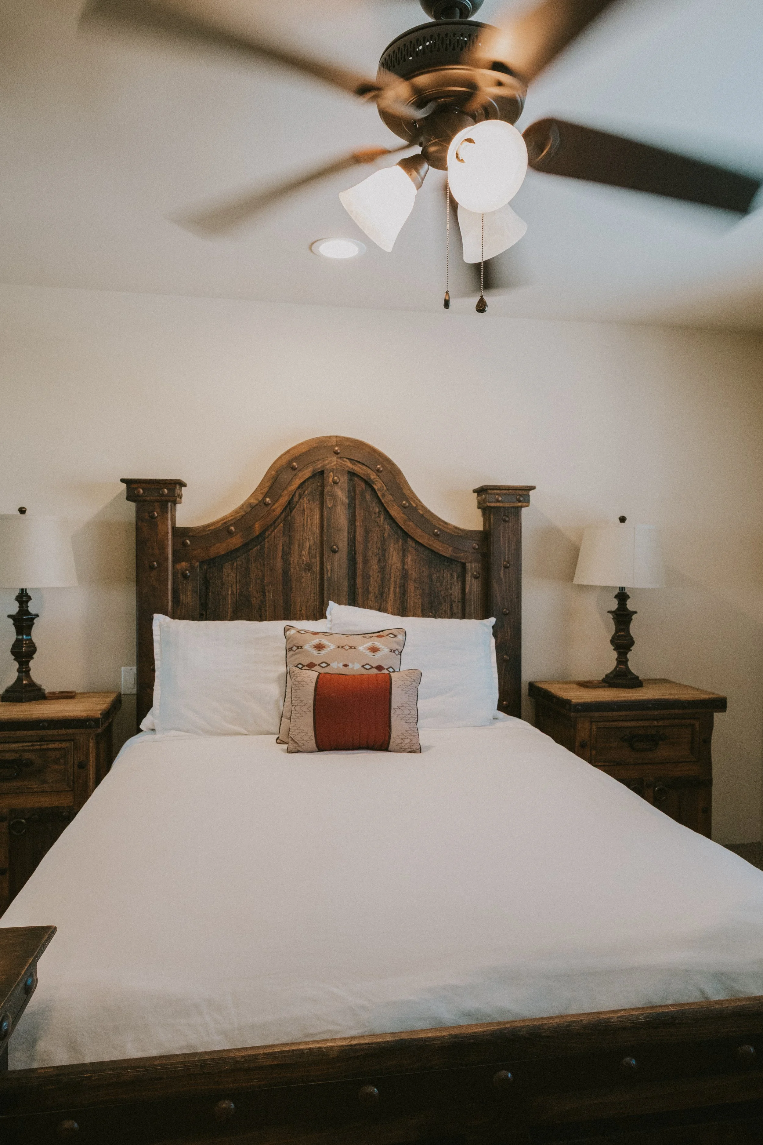 A bedroom featuring a large wooden bed with a white bedspread, a decorative pillow, and a wooden headboard. There are matching nightstands with lamps on each side of the bed. The ceiling has a fan with attached light fixtures and pull chains.