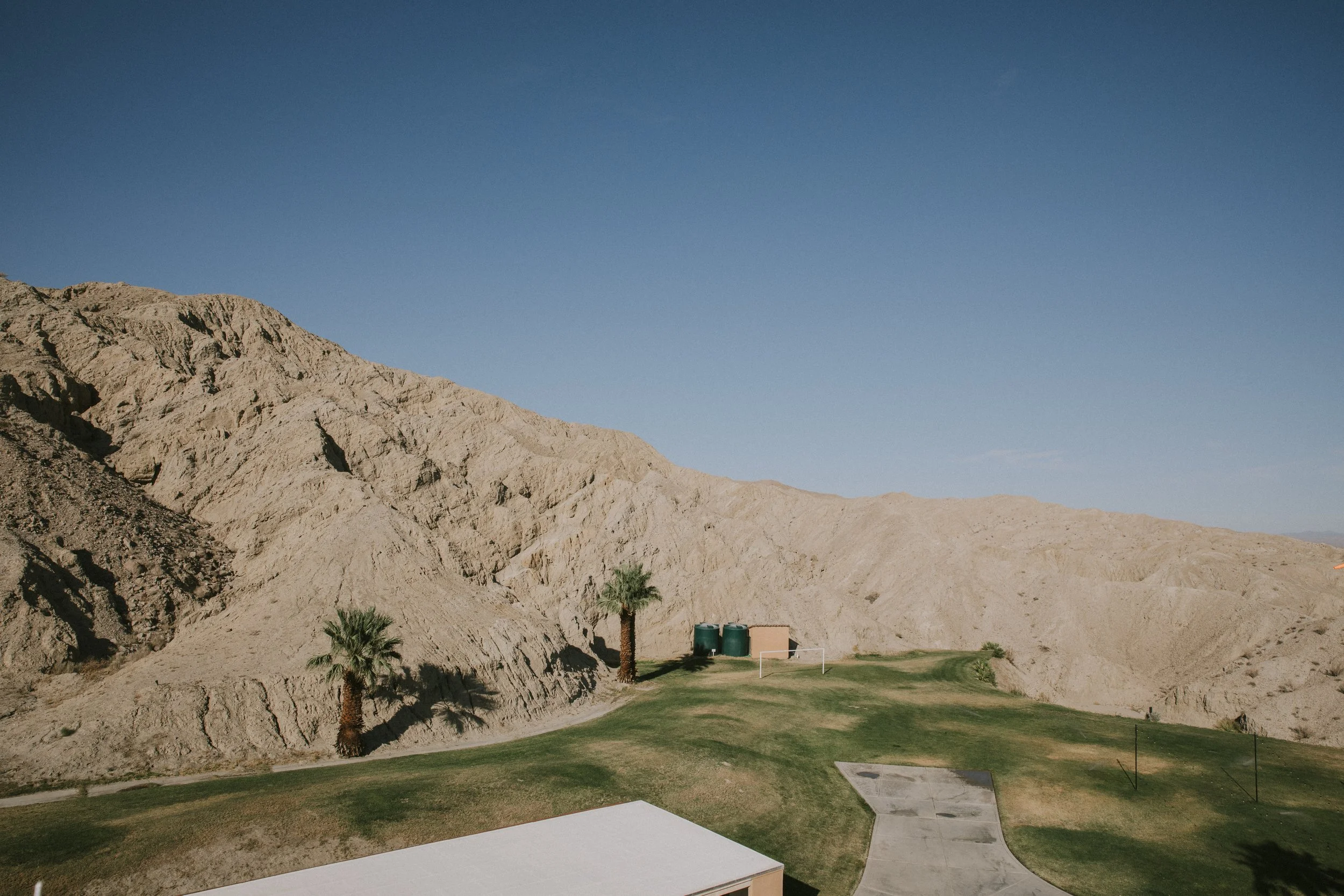 Desert mountains with a small backyard in the foreground that includes green grass, a few palm trees, a soccer goal, and concrete pathways.