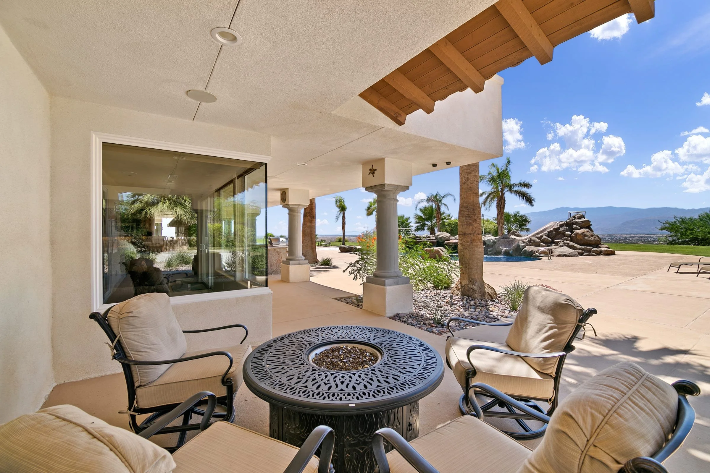 Patio with four cushioned chairs and a decorative round metal table in a backyard. Paved ground, some palm trees, a rock formation with waterfall, and mountain view in the background.