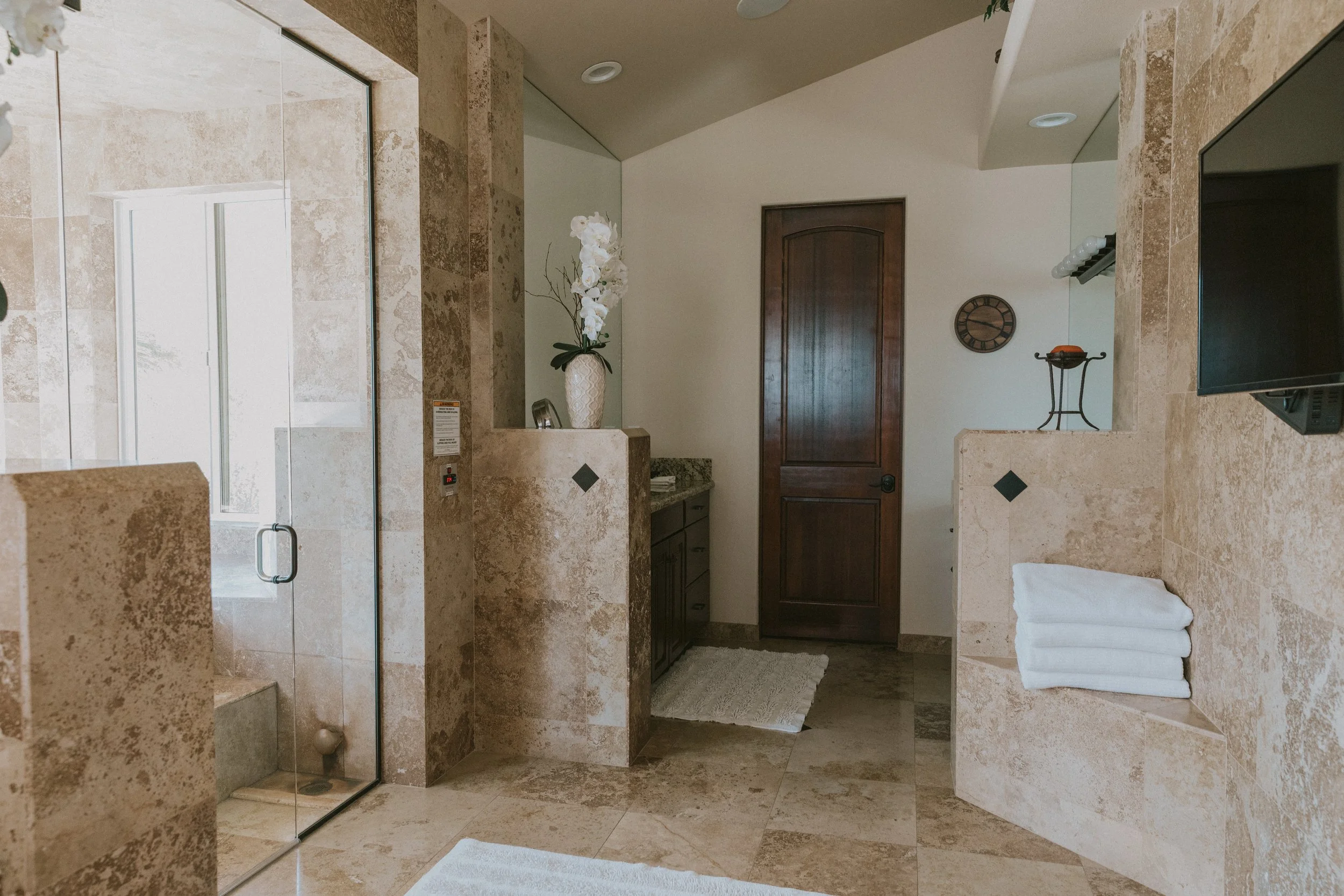 Bathroom with beige tiled walls and floor, frosted glass shower door, dark wood cabinet, wall clock, decorative towel rack with towels, potted plant on vanity, wall-mounted TV, and beige rug.