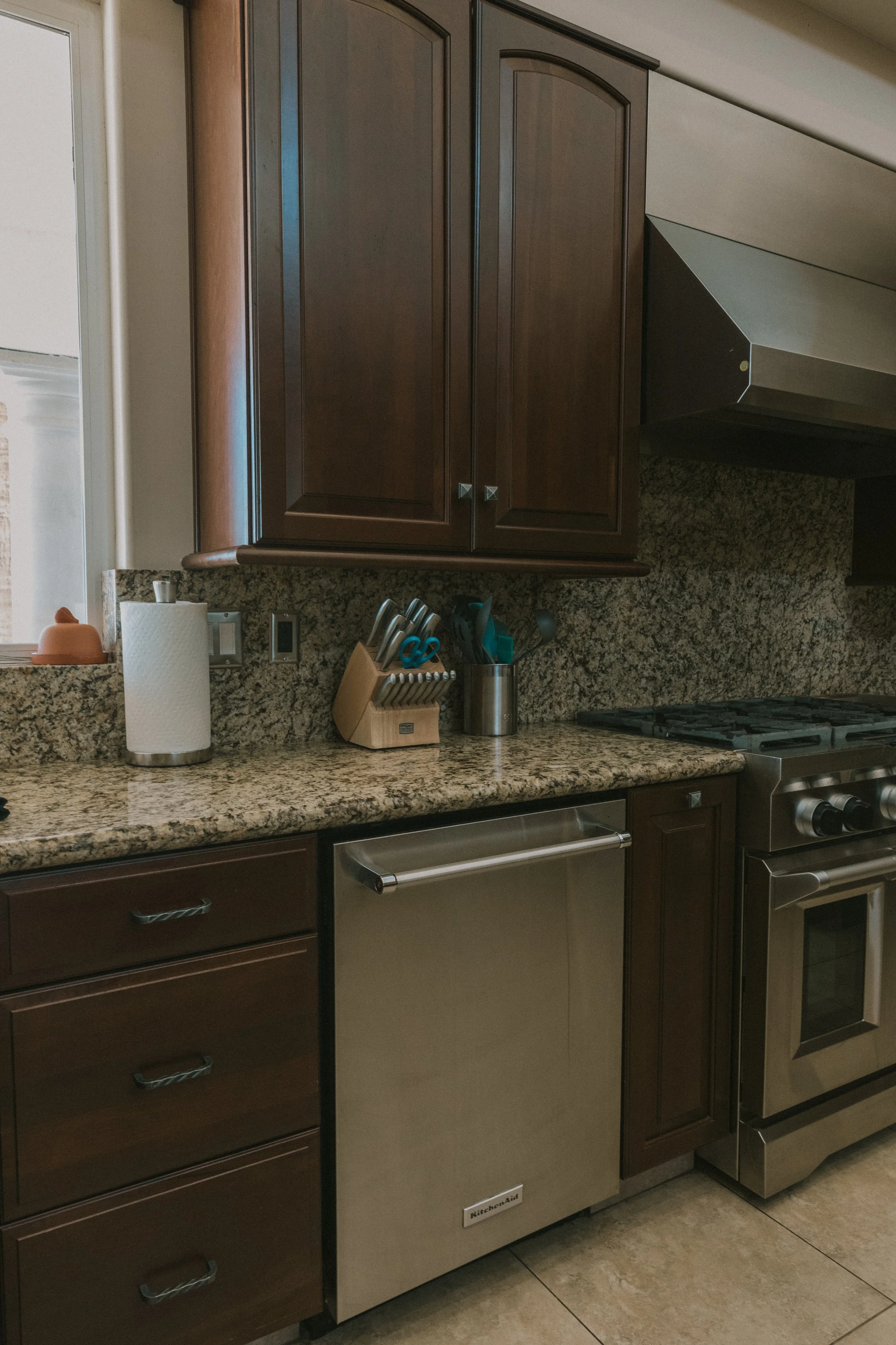 A kitchen with dark wood cabinets, granite countertops, a stainless steel dishwasher, a paper towel roll, and a knife block on the counter.