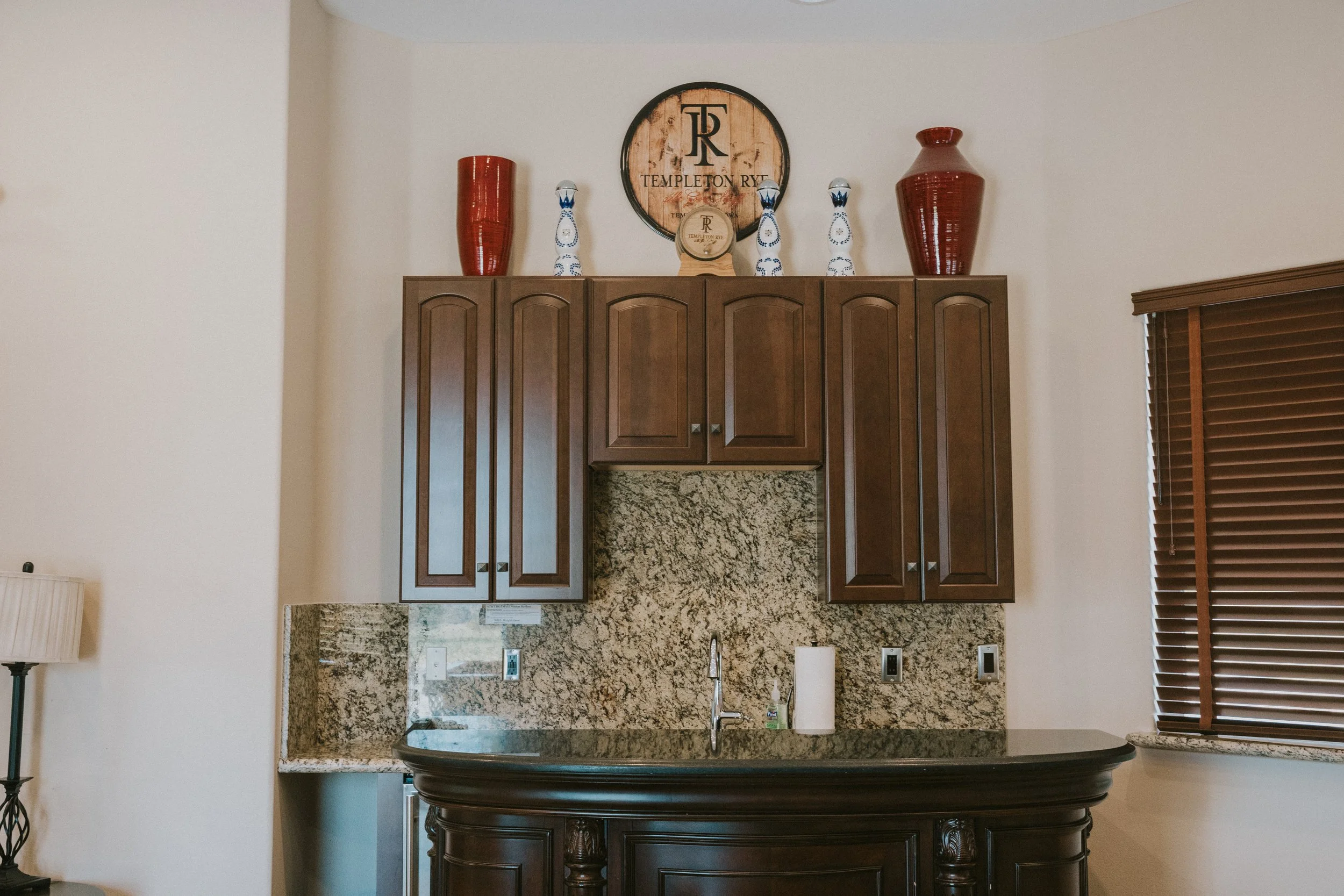 A kitchen with wooden cabinets, granite countertops, a stainless steel sink, and decorative vases and a sign on top of the cabinets.