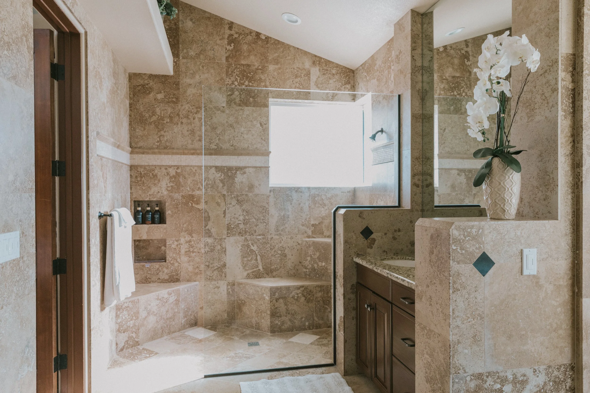Bathroom with beige marble tiles, a large shower area with a glass door, a window, a vanity with dark wood cabinets, a sink, a orchid plant in a decorative vase, and a towel hanging on a wall hook.