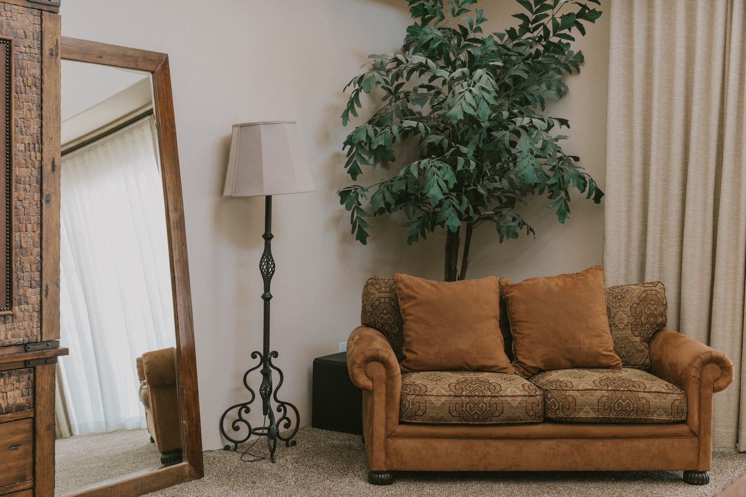A living room corner with a large potted plant, a wooden framed mirror, a floor lamp, a brown patterned couch with orange pillows, and beige curtains.