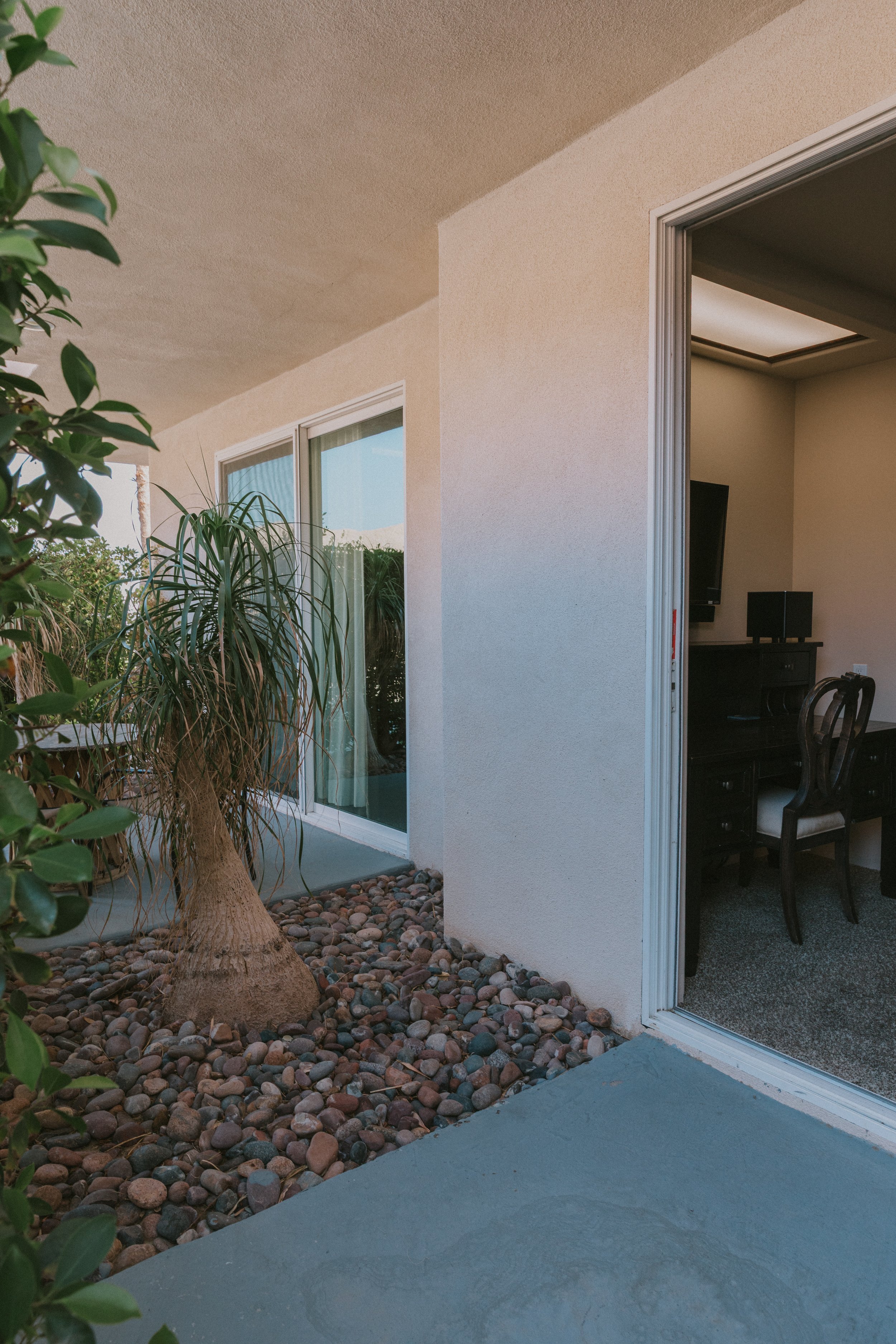 View of an indoor room with a desk, radiator, black tv, and chair through an open sliding door, with an outdoor patio area featuring a potted plant, window, and pebble ground.