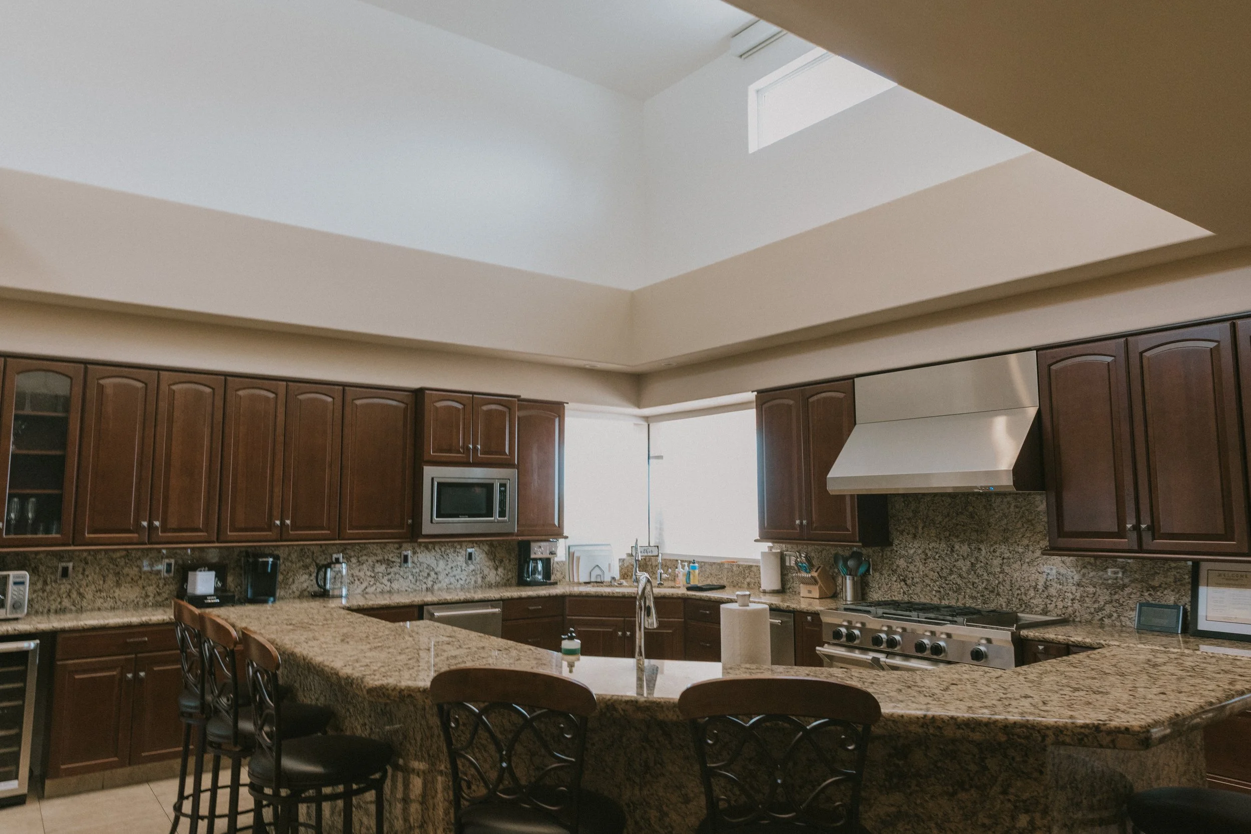 A spacious kitchen with dark wooden cabinets, granite countertops, a kitchen island with seating, stainless steel appliances, and a skylight overhead.