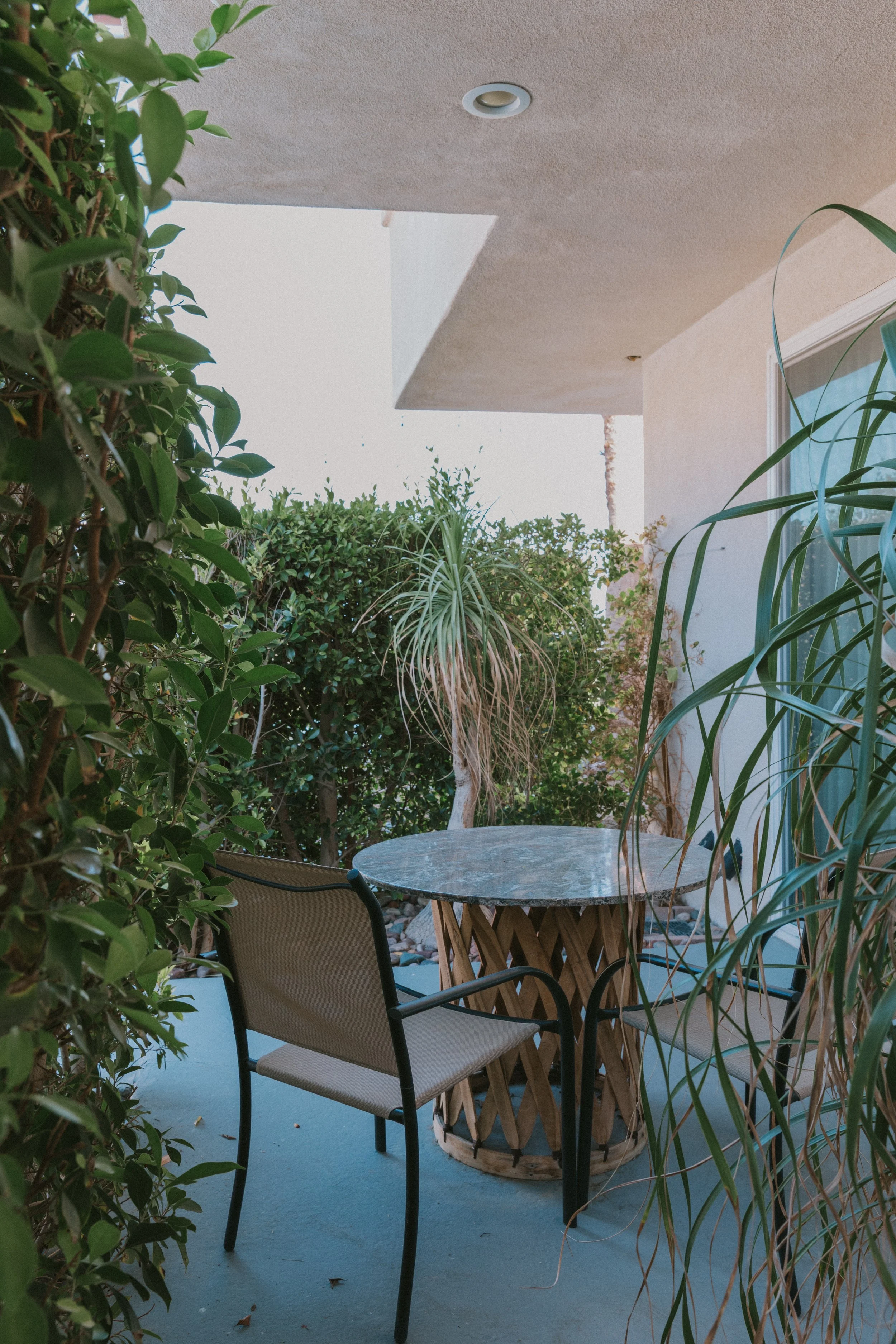 A patio with a round marble-top table surrounded by two beige chairs, lush green plants, and a beige wall with a window, under a ceiling with a recessed light. The patio floor is painted blue.