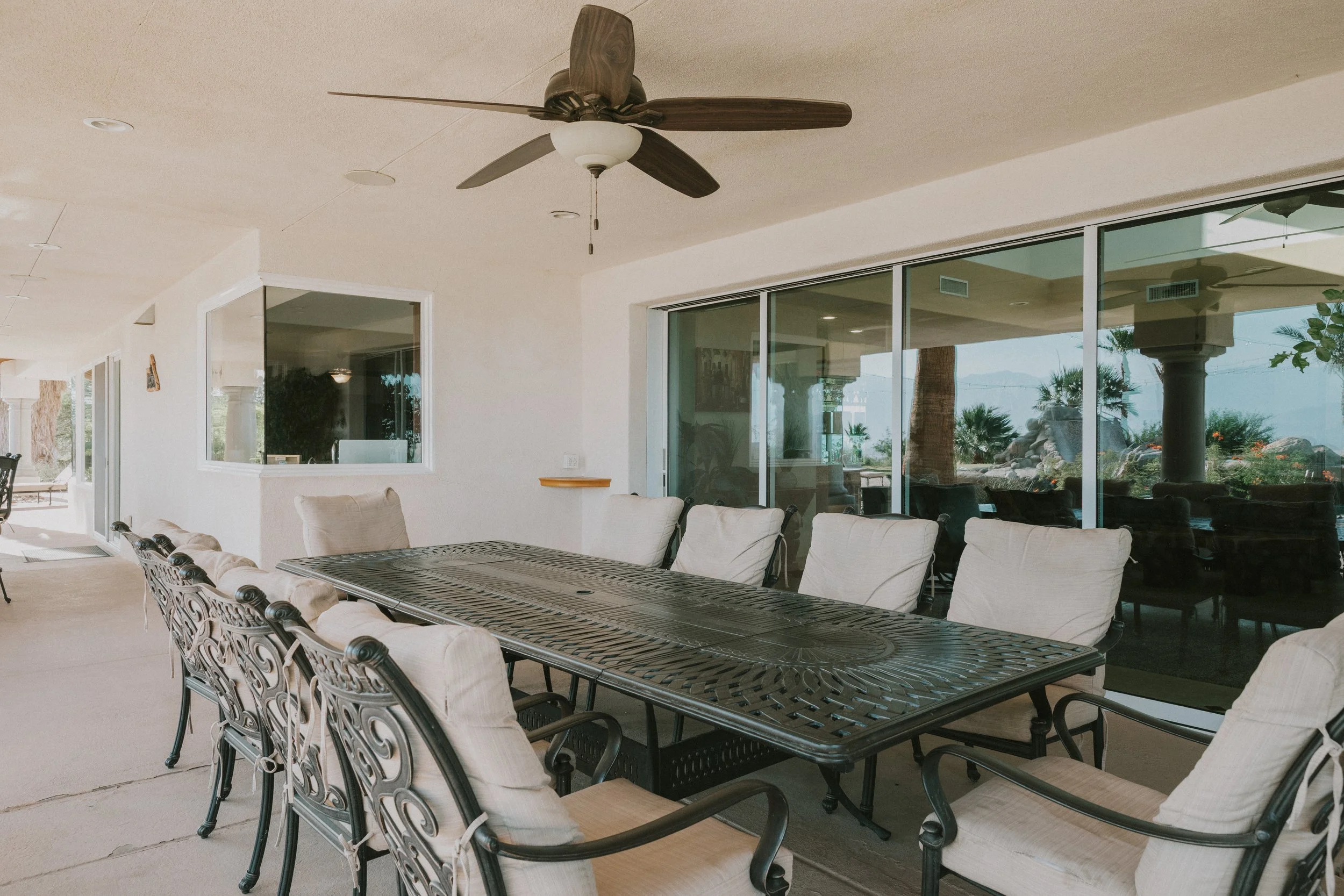 Covered outdoor patio with a large black metal dining table and light-colored cushioned chairs, ceiling fan, and large sliding glass doors leading outside.