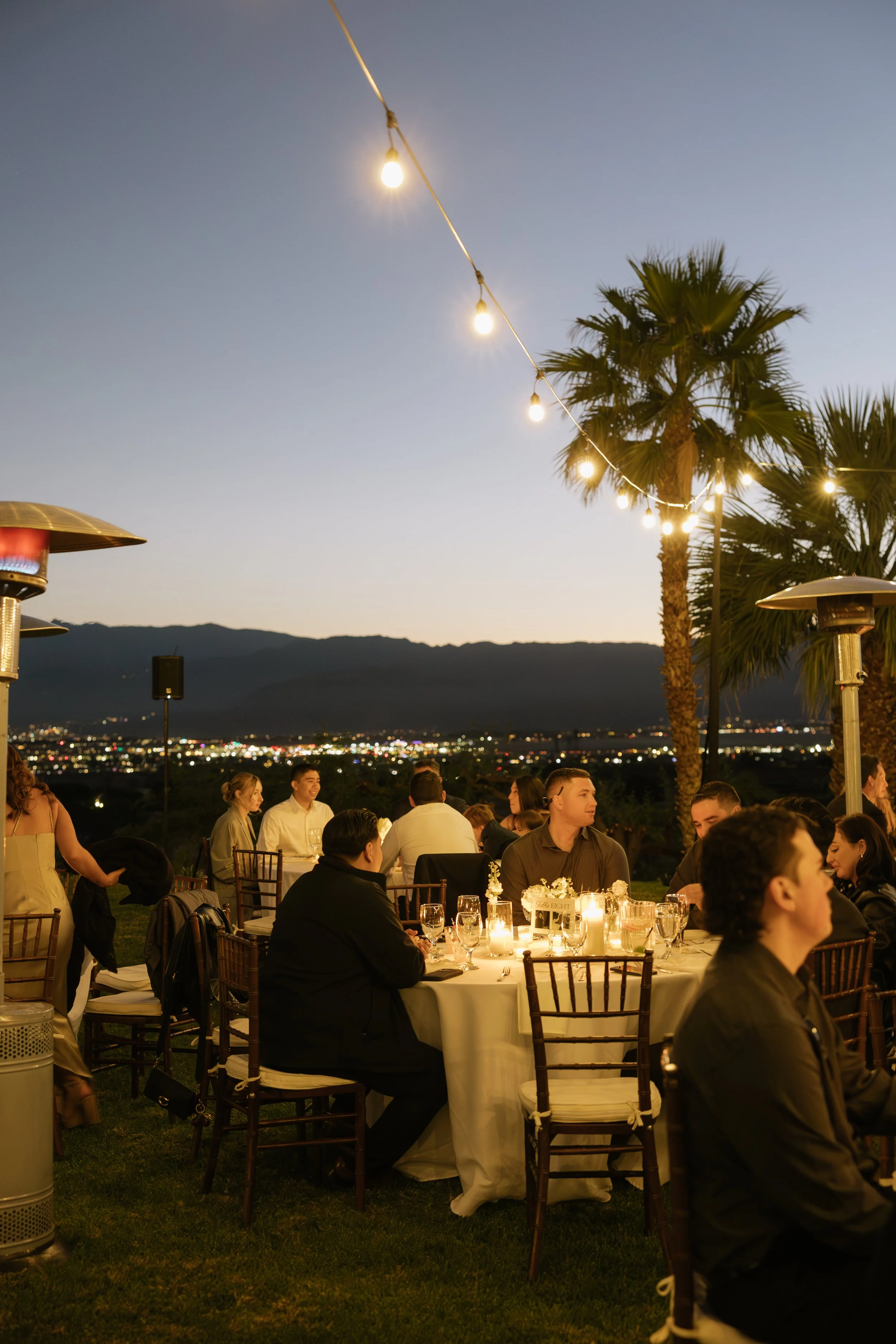 An outdoor evening celebration with people sitting at tables under string lights and palm trees, overlooking a cityscape and mountains in the distance.