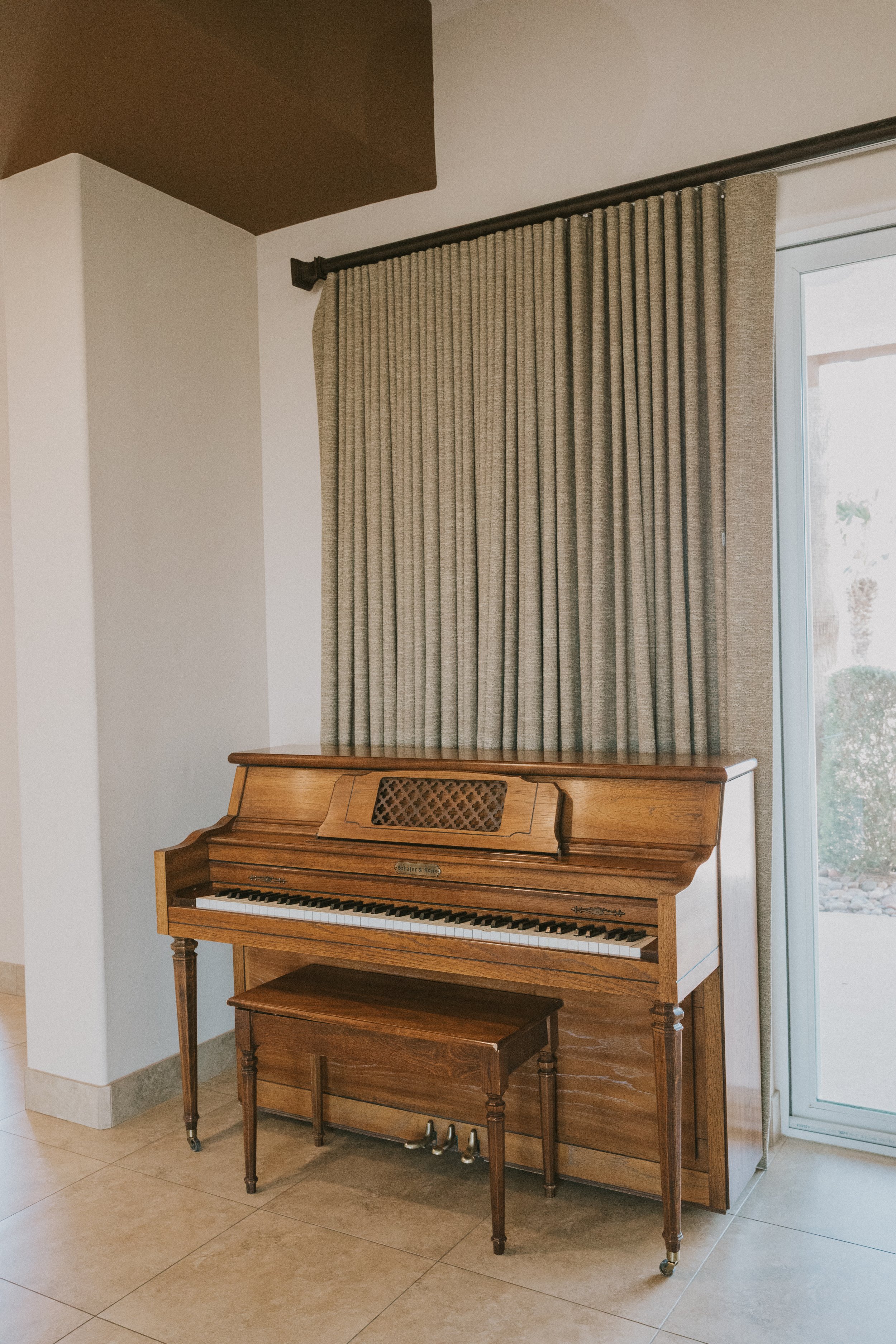 An upright wooden piano with a matching bench in front of a window with beige curtains, located in a room with tiled floor and white walls.