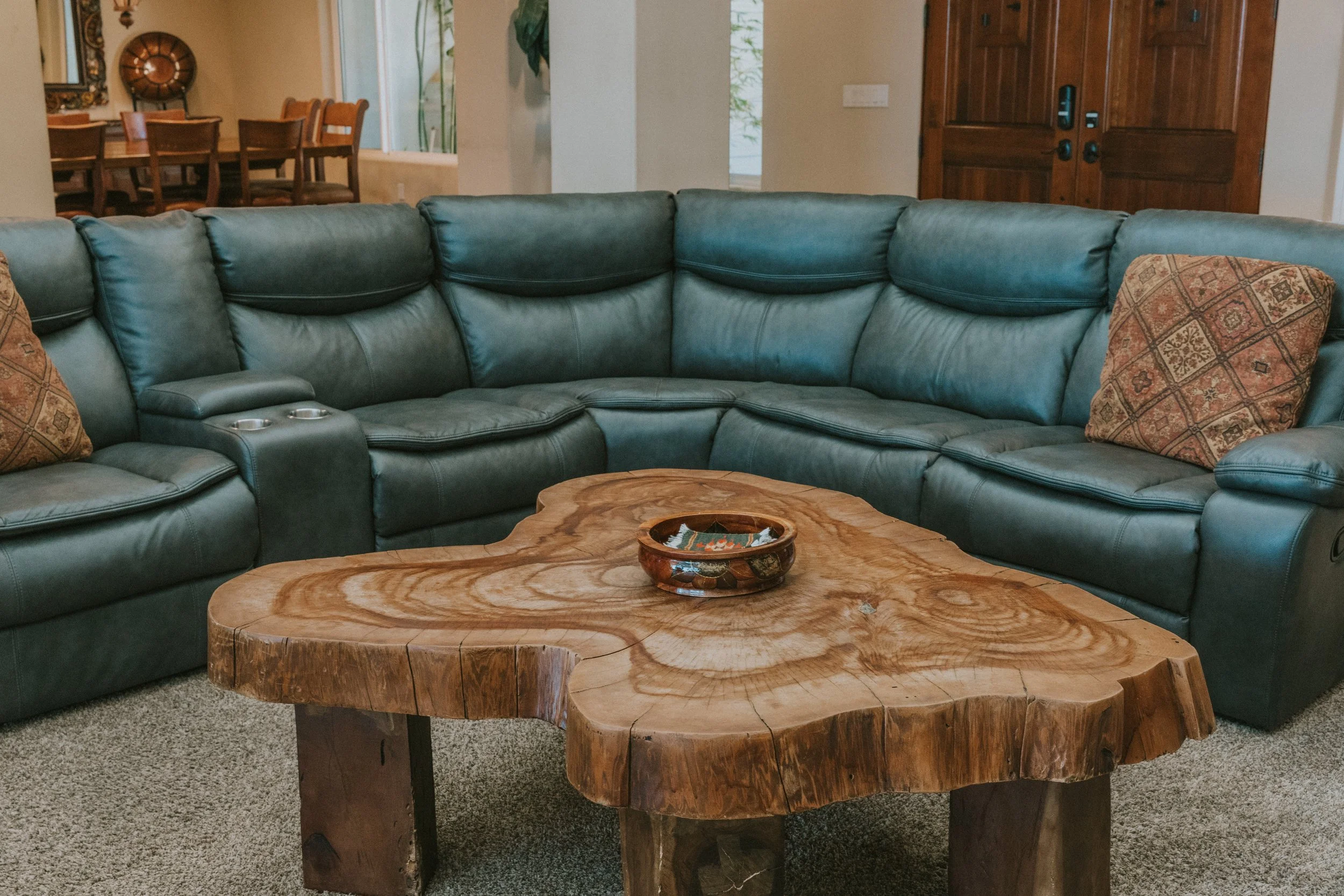 A living room with a corner sectional leather sofa, beige walls, a wooden coffee table, and decorative pillows. A dining area with a wooden table and chairs is visible in the background.