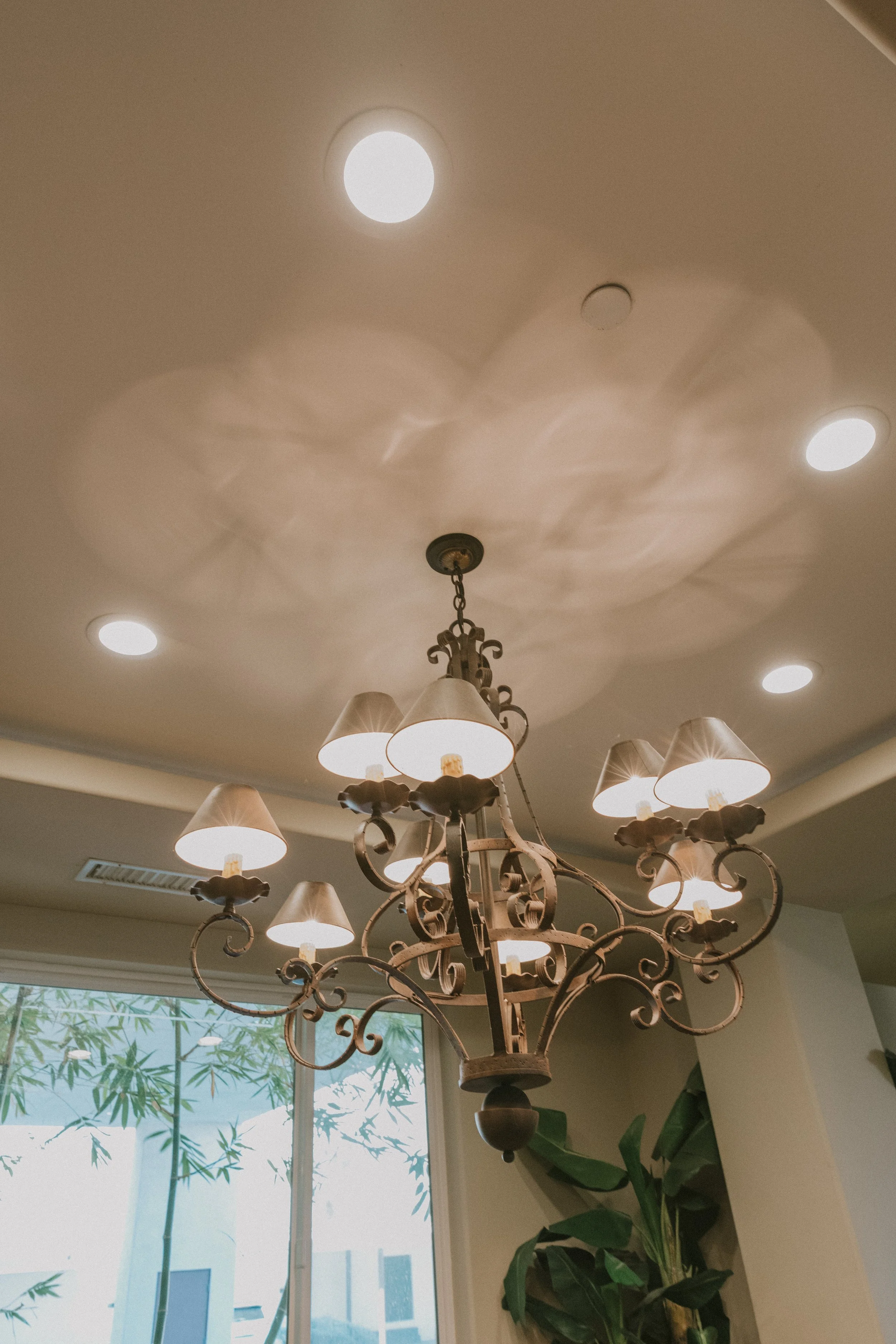 An ornate chandelier hanging from a ceiling with five bright ceiling lights and a central light fixture, in a room with a large window and green plants outside.
