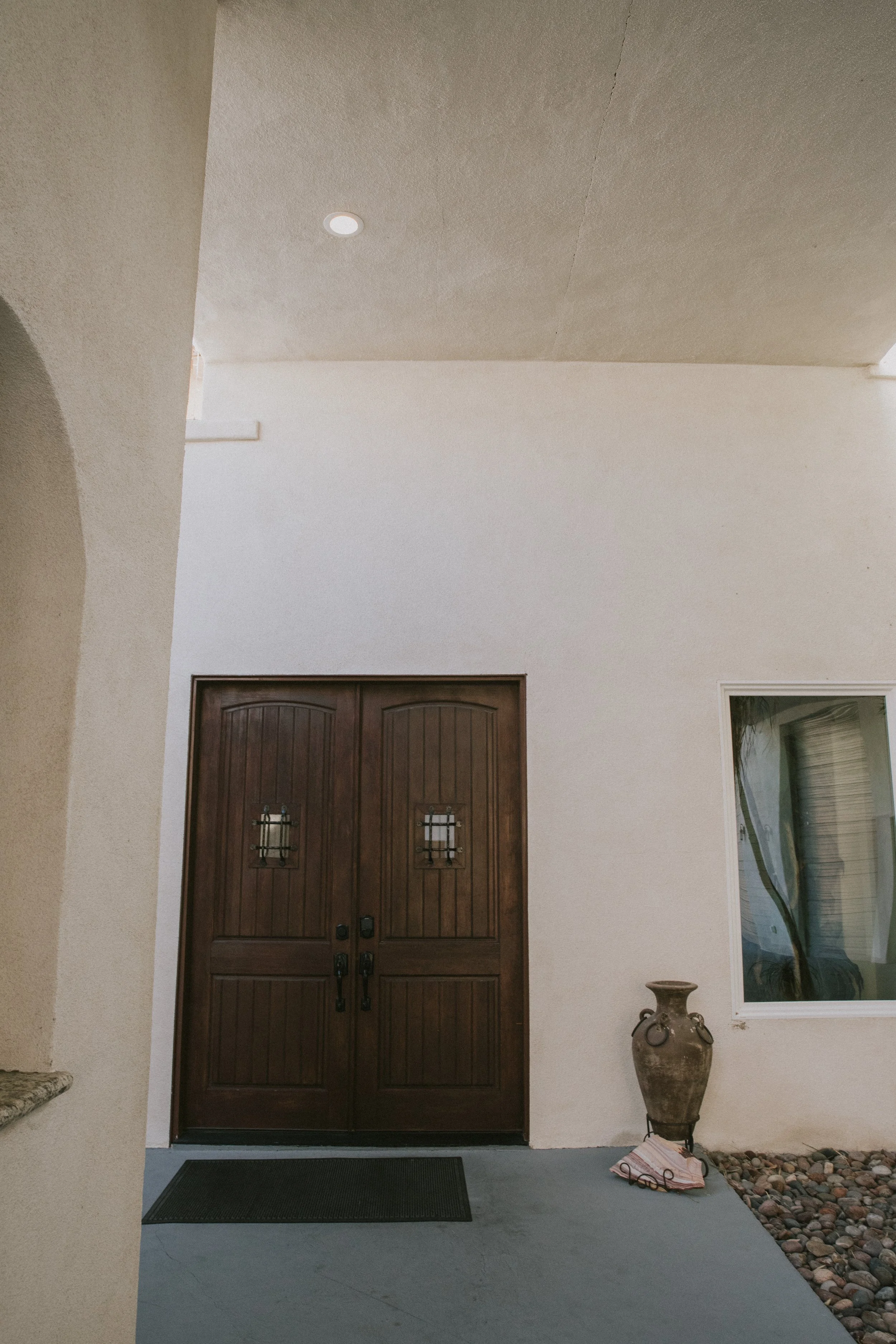 Exterior view of a house front entrance with dark wooden double doors, a beige adobe-style exterior wall, a large window with a reflection, and a decorative vase on a stand next to the door on a bed of small rocks.