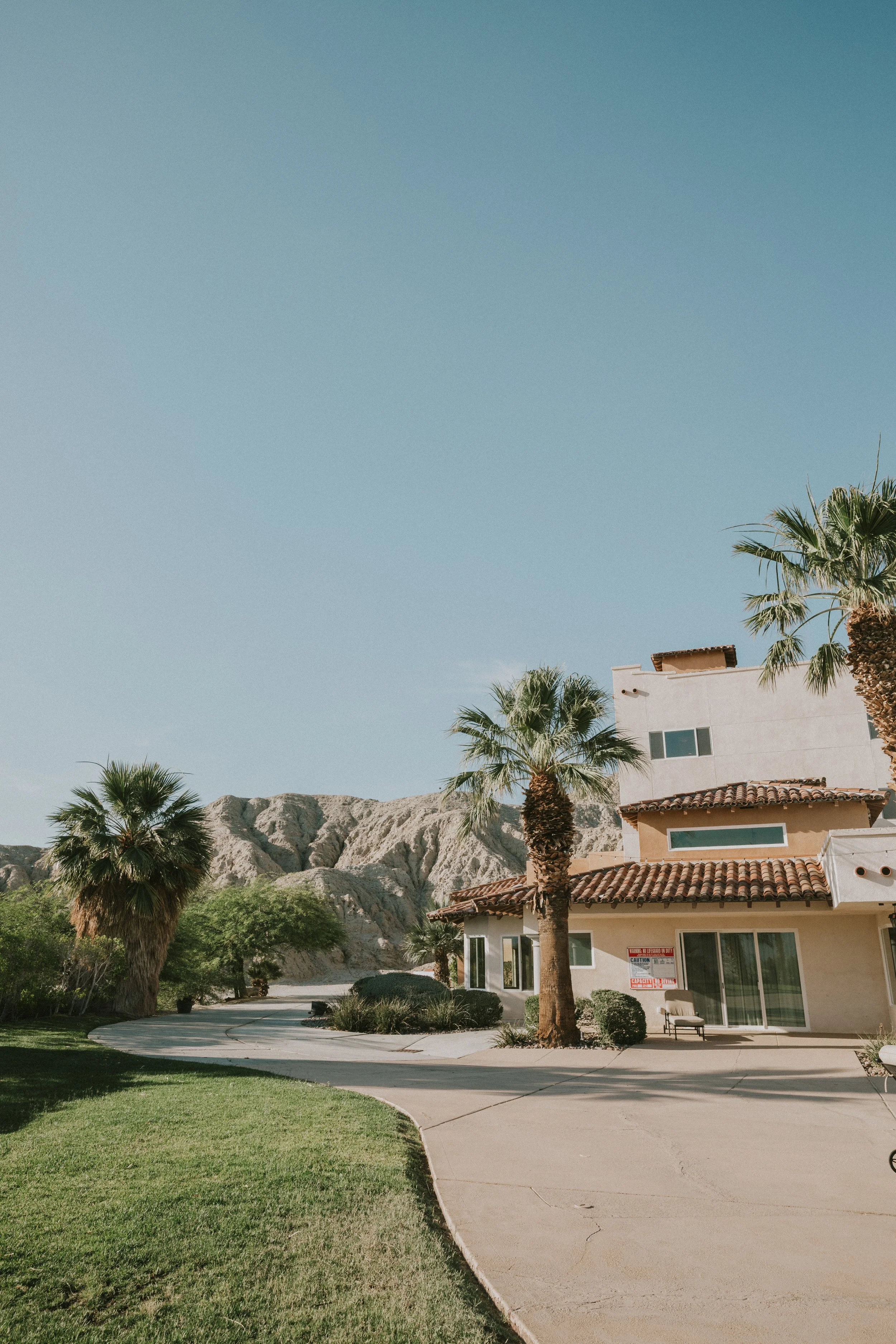 A southwestern style house with a red-tile roof and white stucco walls, surrounded by palm trees, with mountains in the background under a clear blue sky.