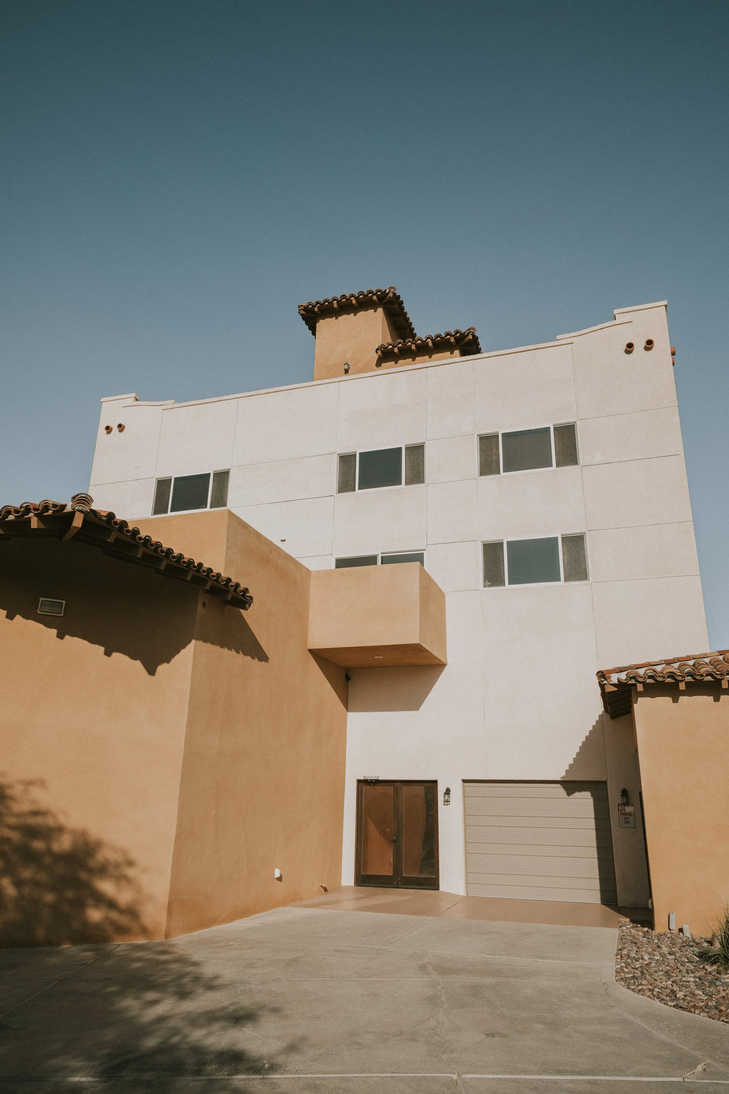 The image depicts a modern multi-story building with a beige and white exterior, featuring small windows, a garage door, and a glass door entrance. The building has a minimalist design with a curved roofline and a small decorative wall extension. The