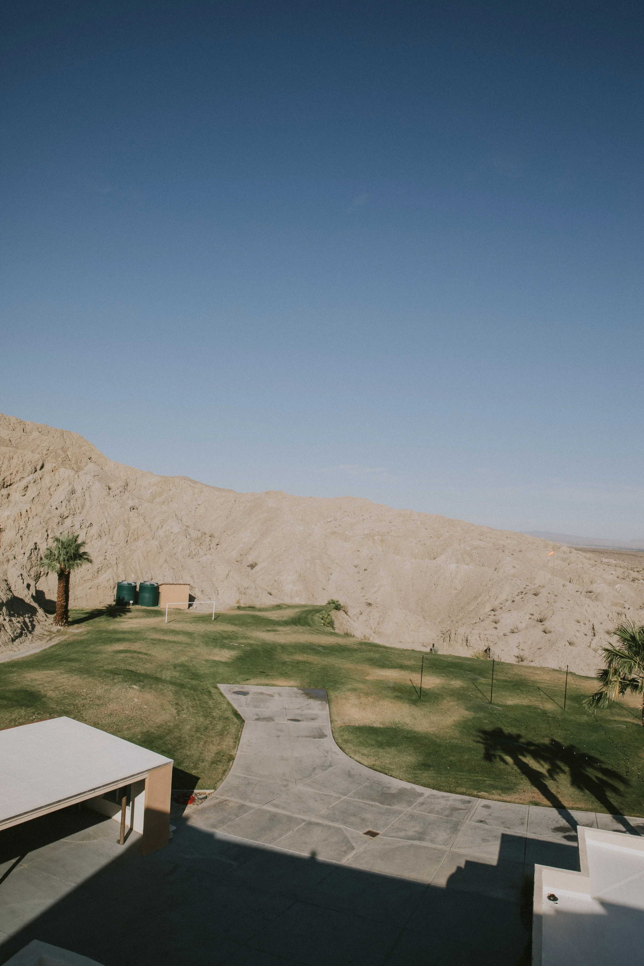 View of a landscaped yard with green grass, a single palm tree, and a concrete walkway in a desert-like mountainous area under a clear blue sky.