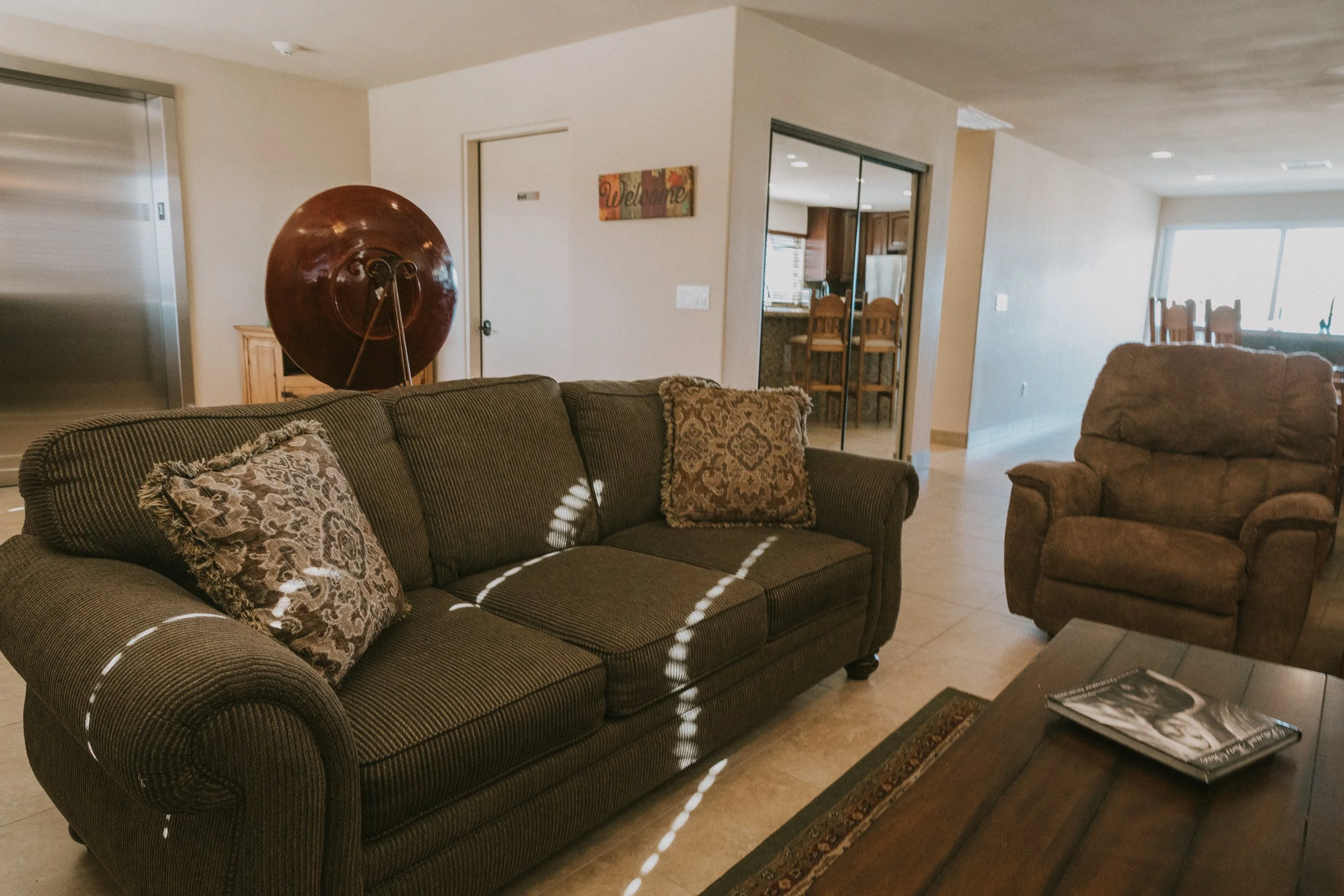 Living room with brown upholstered sofa, patterned throw pillows, a matching armchair, coffee table with magazines, wood flooring, and sliding glass doors leading outside. An elevator and a welcome sign are visible in the background.