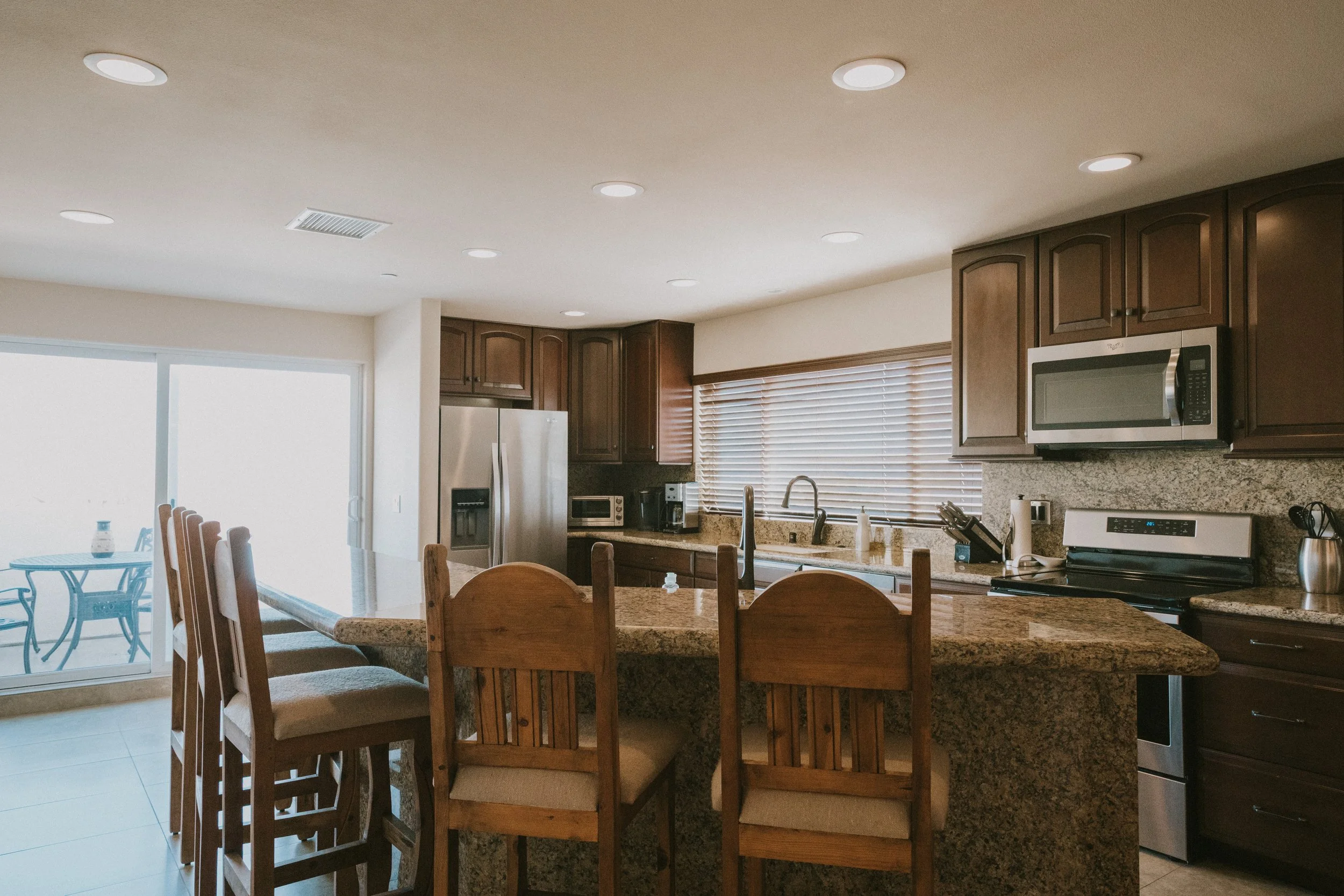 A spacious kitchen with wooden cabinets, a granite island, stainless steel appliances, and large windows with blinds, illuminated by ceiling lights.