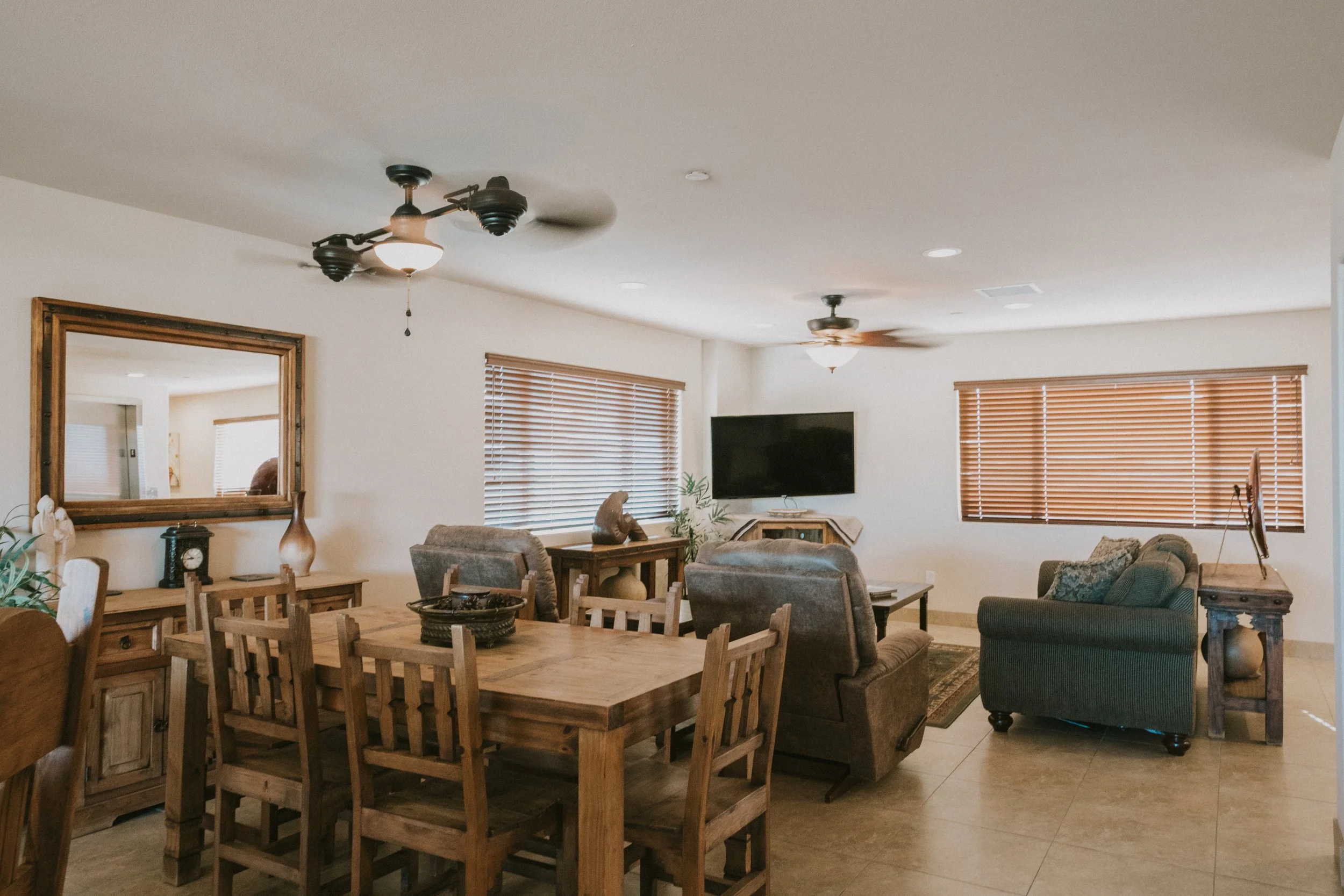 A living room with a wooden dining table and chairs, sofas, a wall-mounted TV, wooden blinds, ceiling fans, and various decorative items.