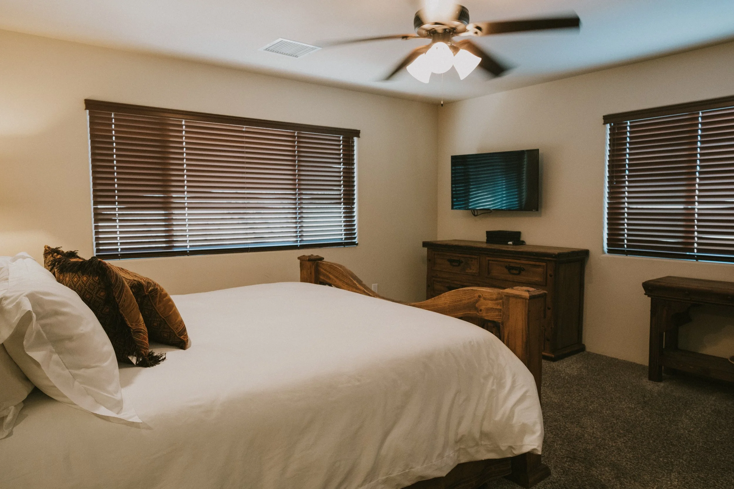A bedroom with a bed, pillows, wooden furniture, window blinds, a wall-mounted TV, and a ceiling fan.