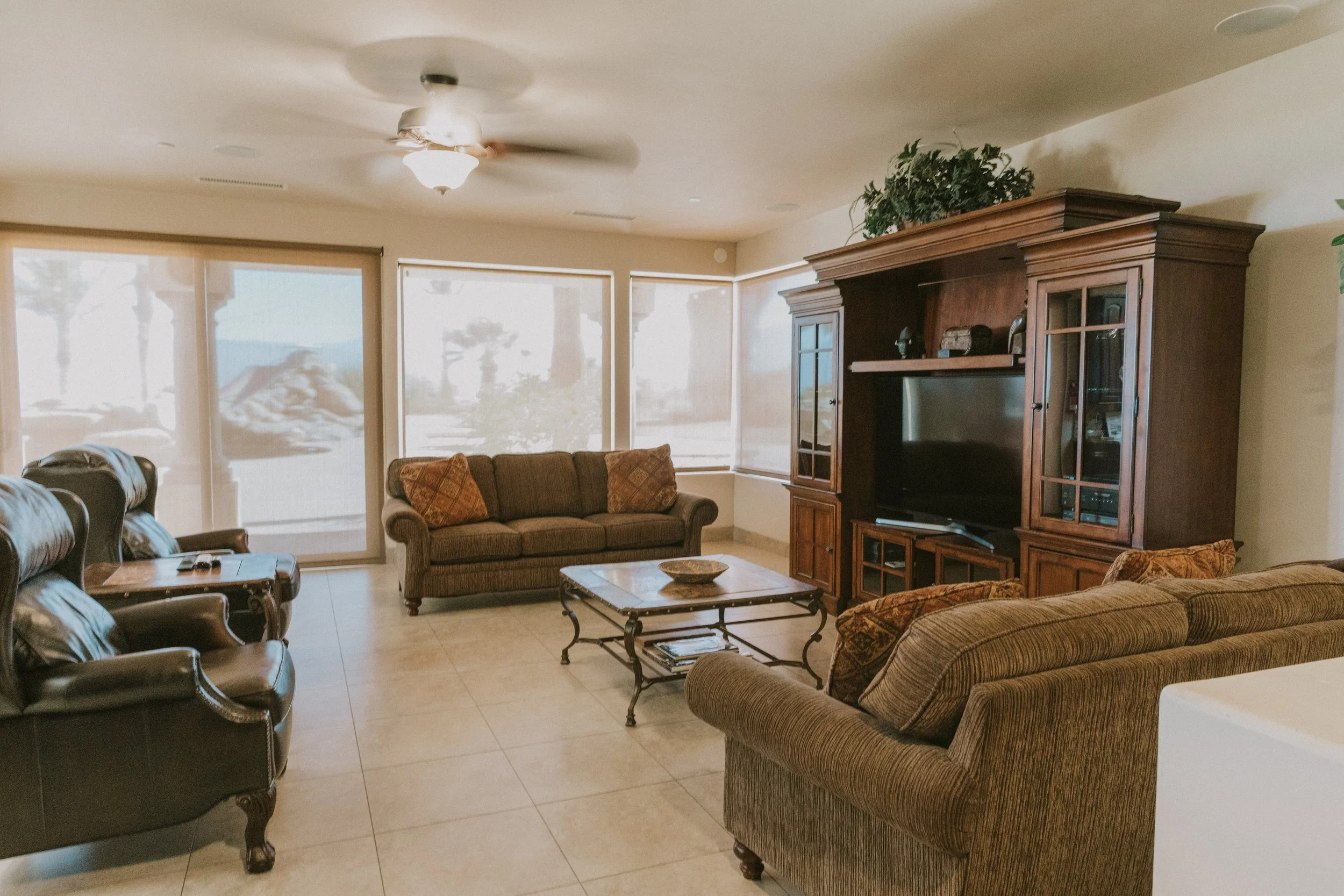 Living room with beige tiled floor, beige walls, large windows with blinds, a ceiling fan, an entertainment center, a sofa with pillows, and leather armchairs.