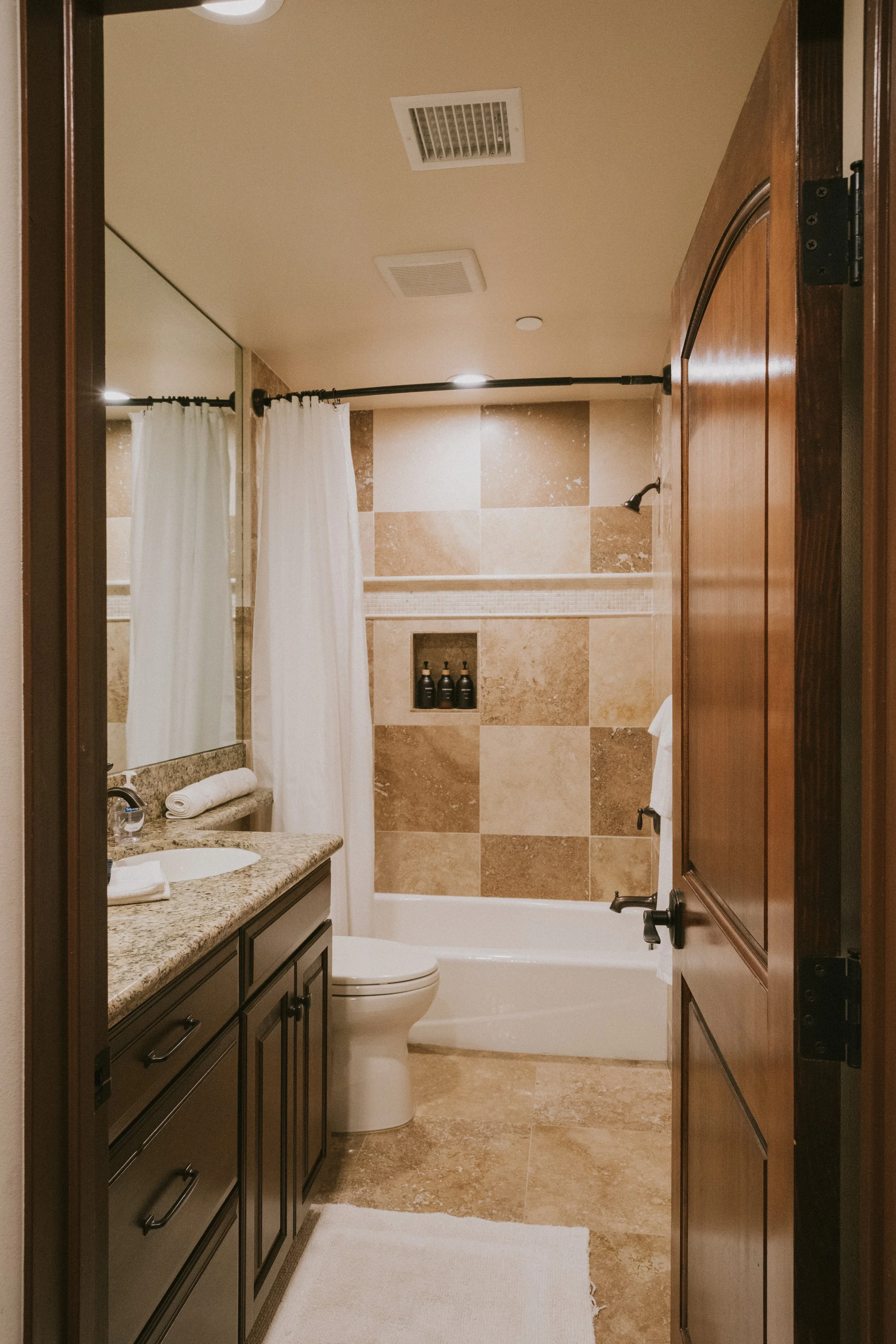 A bathroom with a dark wooden door, granite countertop vanity, oval sink, toilet, bathtub with shower, shower curtain, and tiled walls in beige and brown tones. There is a large mirror above the vanity and black fixtures. A white bath mat is on the f